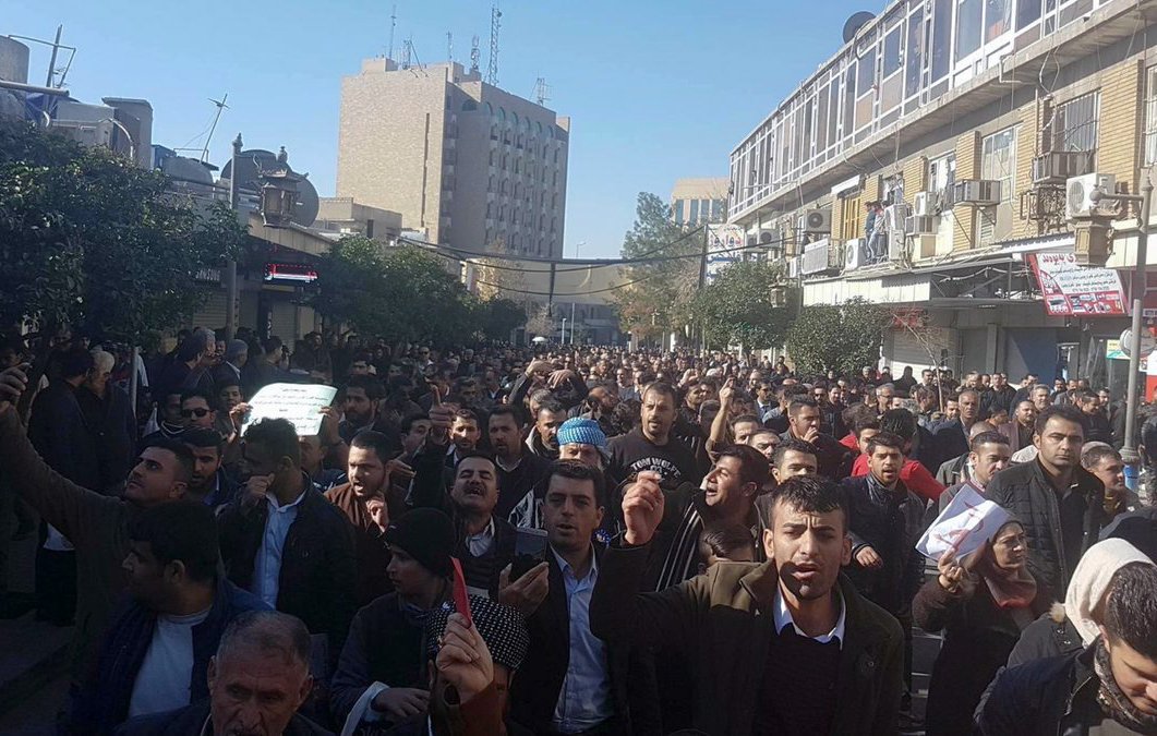 L'image montre une foule dense de personnes rassemblées dans une rue, manifestant visiblement. Les gens tiennent des pancartes et semblent exprimer des revendications. En arrière-plan, on peut voir des bâtiments urbains, ce qui indique que cela se déroule dans une ville. L'atmosphère semble énergique, avec des manifestants qui lèvent les bras et chantent. Les manifestants portent des vêtements variés, et on peut apprécier la diversité des participants.