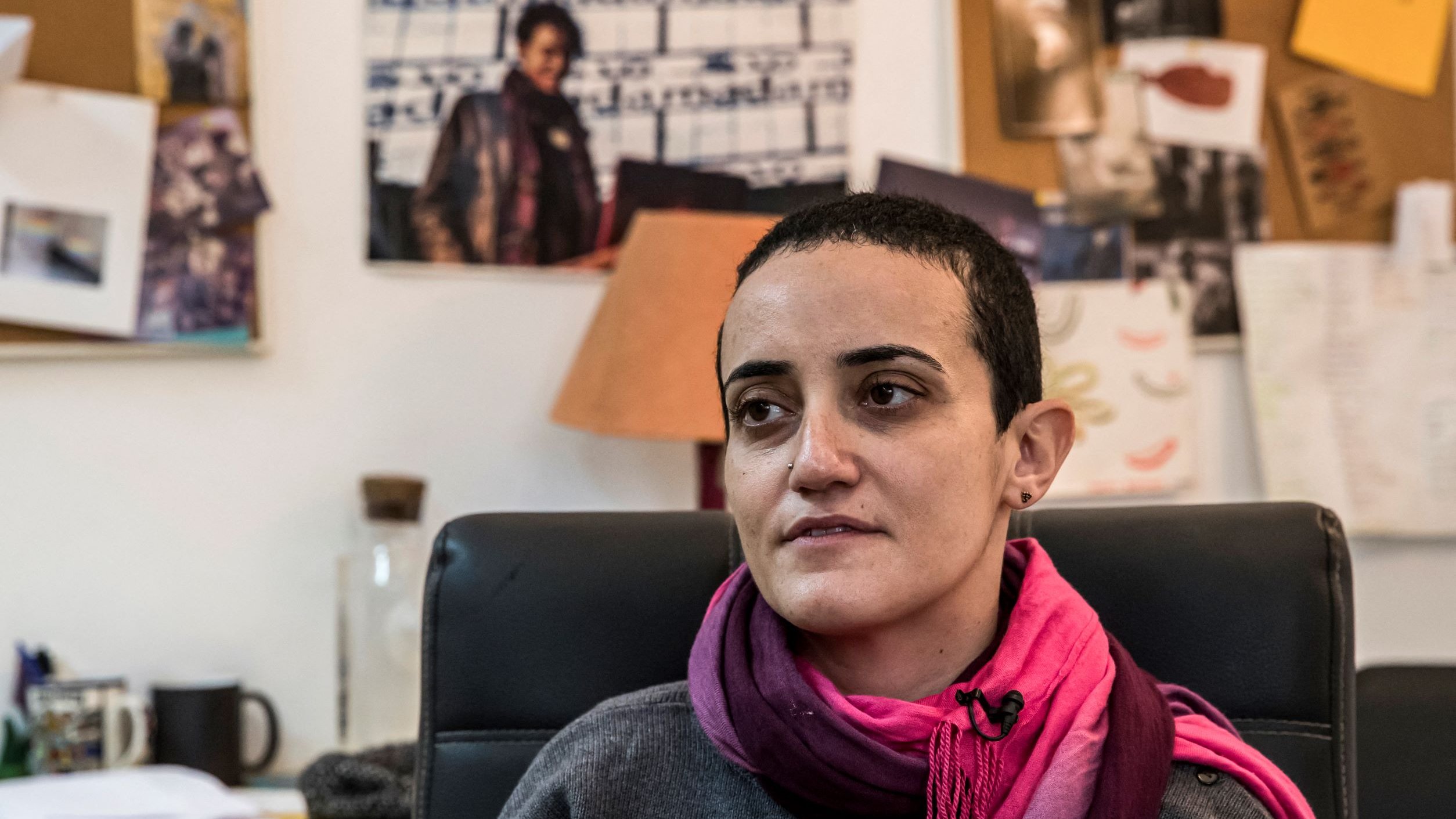 تظهر الصورة امرأة في مكتب، حيث تضع على كتفيها وشاحًا ورديًا. تصفح عينيها بعمق، ويبدو أنها تفكر أو تعبر عن مشاعر معينة. في الخلفية، يمكن رؤية مجموعة من الأوراق والصور المعلقة على الحائط، بالإضافة إلى أضواء وزرع، مما يعطي انطباعًا عن بيئة العمل أو الإبداع. الأجواء تبدو غير رسمية، وتعكس شخصية المرأة المتواجدة في الصورة.