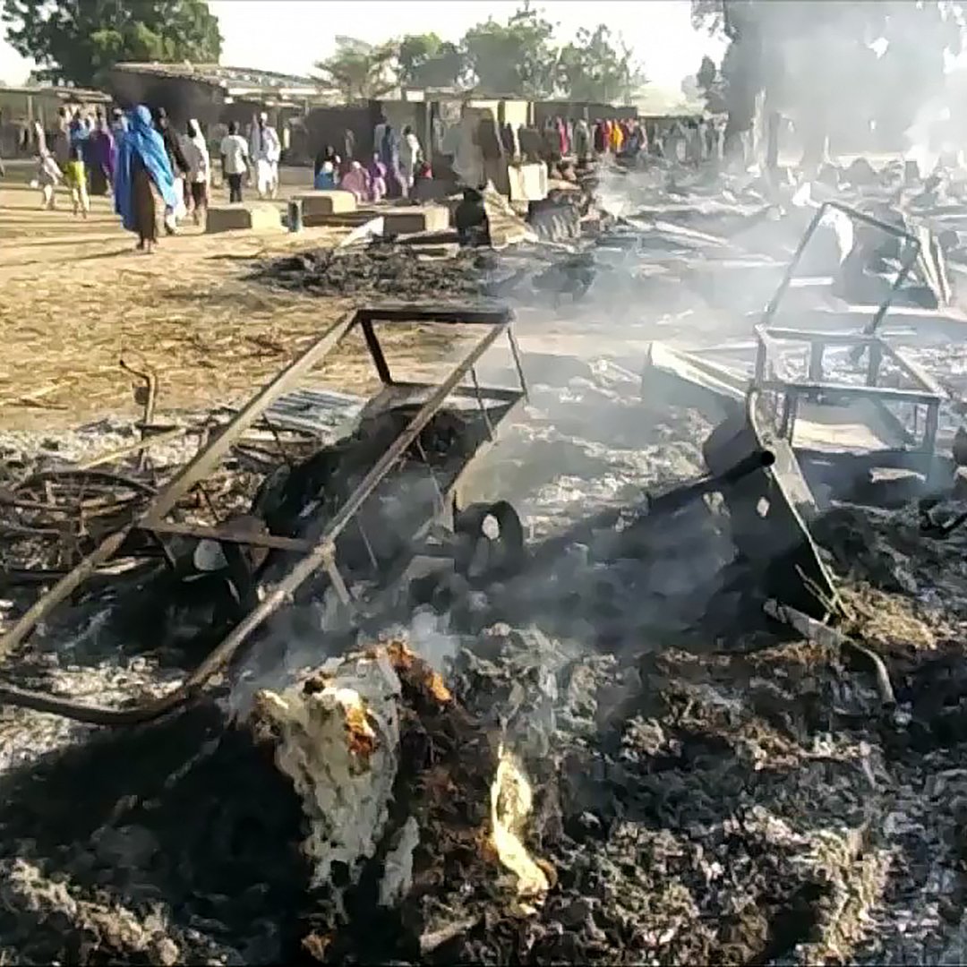 تظهر الصورة مشهدًا من مكان تعرض لحريق، حيث توجد بقايا هياكل معدنية متفحمة ومليئة بالدخان. الأرض مغطاة بالفحم والرماد، مما يدل على اشتعال النار مؤخرًا. يمكن رؤية بعض الناس في الخلفية، مما يشير إلى أنهم ربما كانوا يتجولون في المنطقة المحيطة. الجو يبدو مدخنًا وغير واضح بسبب الدخان المتصاعد.