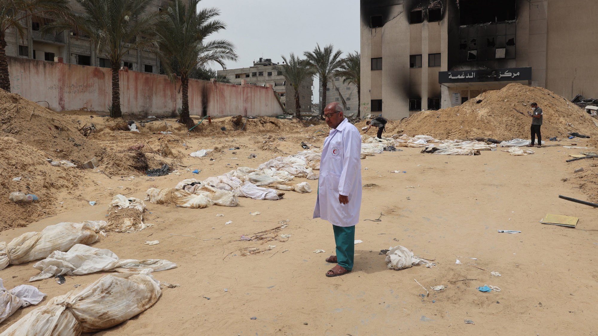 The image depicts a scene in a sandy area where several bodies, covered in white shrouds, are lying on the ground. In the foreground, a man wearing a white coat stands solemnly. In the background, there are damaged buildings, some of which appear to be burned. Palm trees line the area, and the overall atmosphere seems somber and grim, indicating a setting of conflict or disaster.
