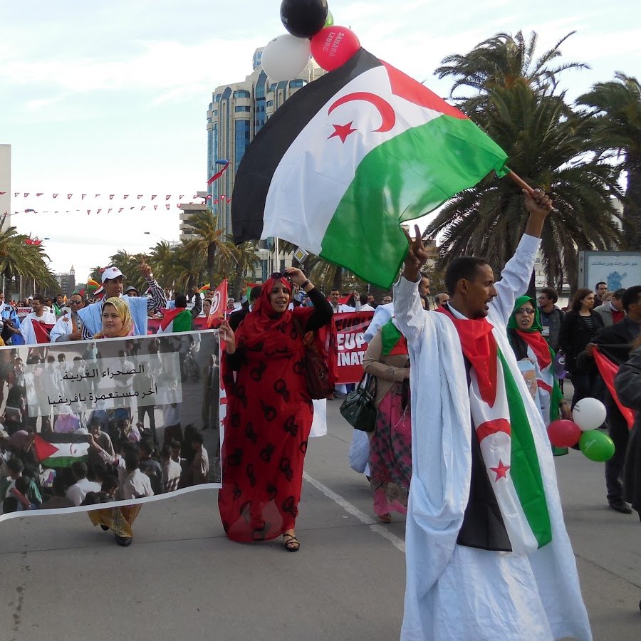 The image depicts a lively demonstration, likely in support of a cause related to Western Sahara. People are marching along a street, holding banners and flags that feature the colors red, green, and black, which are associated with the Sahrawi nationalist movement. The crowd includes both men and women, some wearing traditional clothing. In the background, palm trees and buildings suggest a warm climate, and festive decorations, like balloons, add to the atmosphere of the event. The banner appears to convey a message related to their cause.