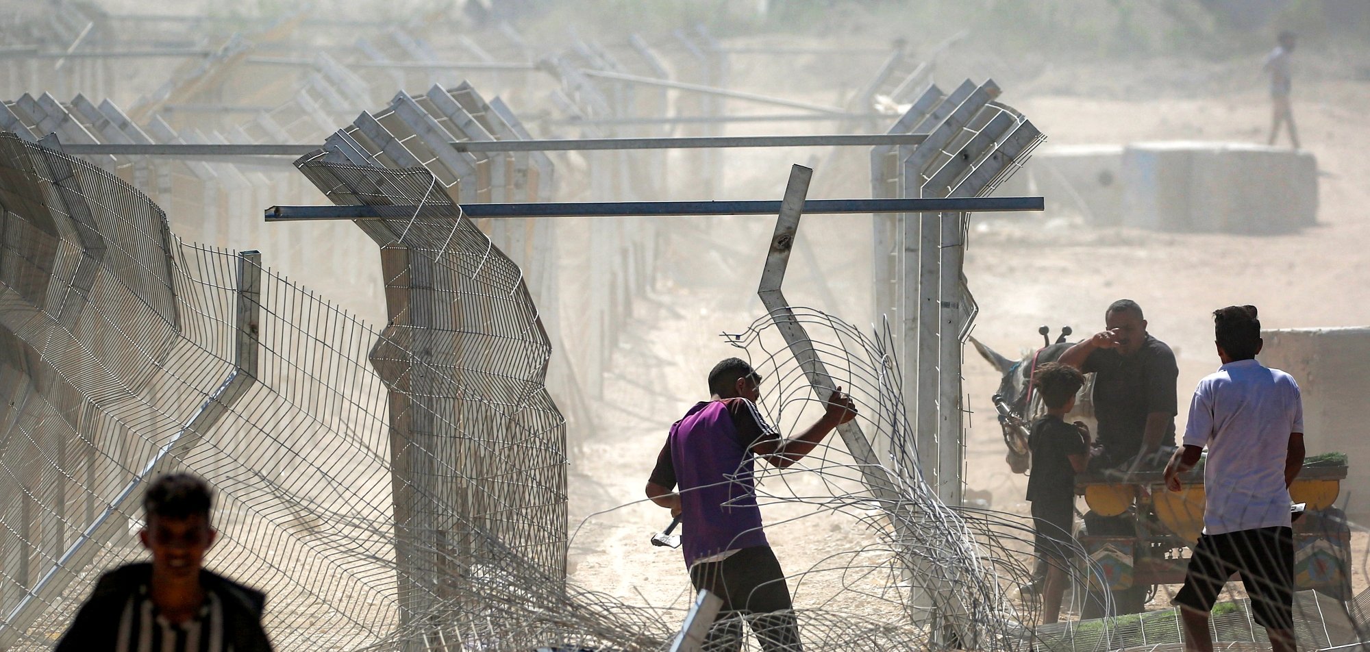 تصویر نشان‌دهنده مردانی است که در حال عبور از حصاری فلزی و برقراری یا تخریب موانع هستند. گرد و غبار در هواست.