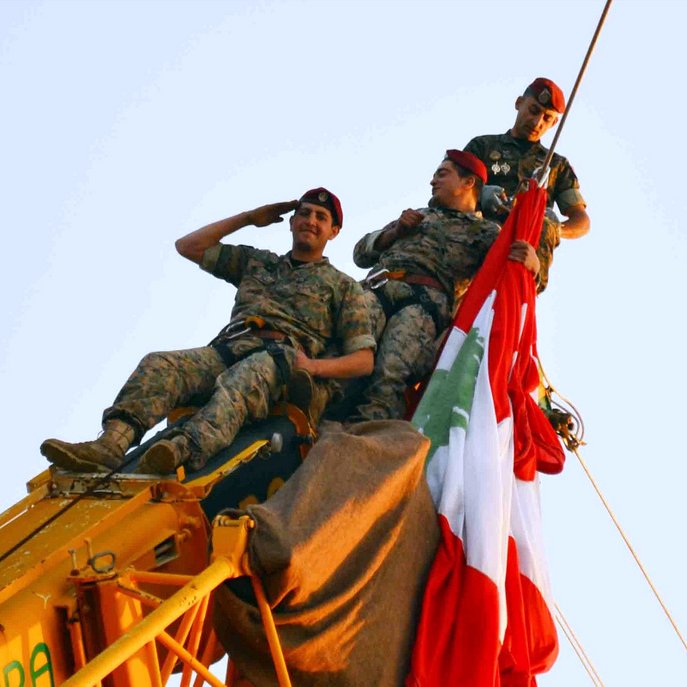 L'image montre trois soldats en uniforme, perchés en haut d'une grue. Ils sont en train de saluer tout en tenant un grand drapeau, probablement celui du Liban. Les soldats affichent un air déterminé et solennel, et l'ambiance semble festive, suggérant une célébration ou un événement important. Le ciel est clair, ce qui met en valeur la scène.