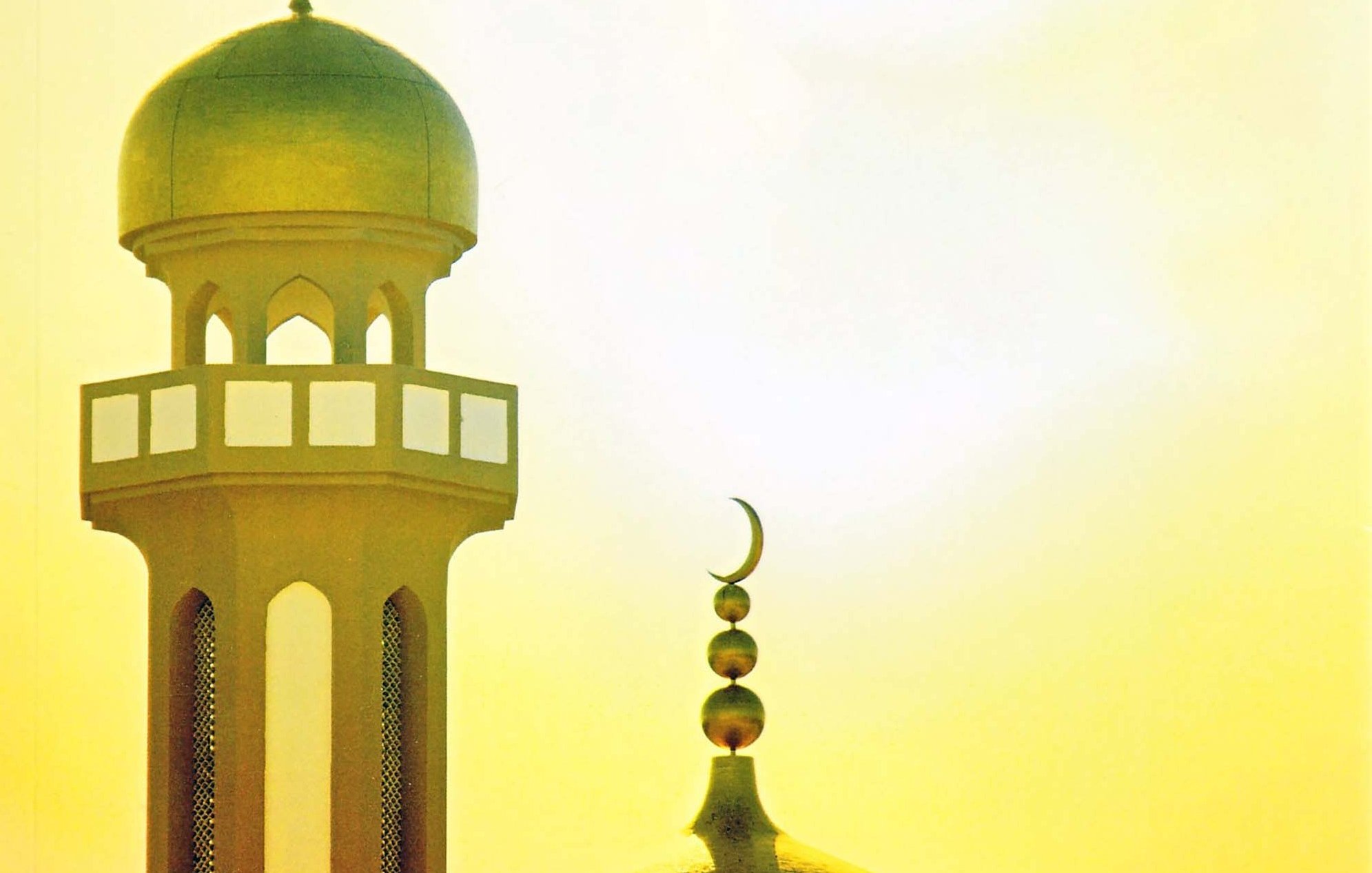 L'immagine mostra il profilo di una moschea, con una cupola grande e una torre alta. La cupola è di forma arrotondata, mentre la torre ha una base decorata con finestre e una punta sormontata da una luna crescente. Sullo sfondo, si intravede un cielo luminoso con tonalità calde di giallo, creando un'atmosfera suggestiva e serena.