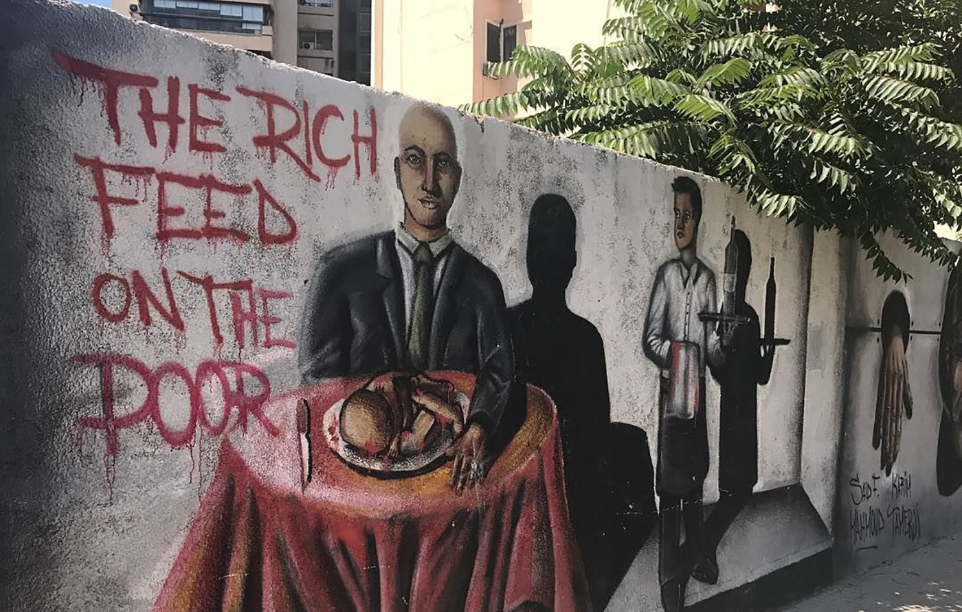 L'image montre une fresque murale colorée sur un mur. Au premier plan, un homme en costume est assis à une table couverte d'une nappe rouge, avec un plat à manger devant lui. À côté, on peut voir une ombre d'un serveur, suggérant un contexte de service alimentaire. Sur le mur, il est écrit "THE RICH FEED ON THE POOR", ce qui évoque un message critique sur les inégalités sociales. Des éléments de l'arrière-plan, comme des bâtiments et de la verdure, ajoutent à l'environnement urbain.