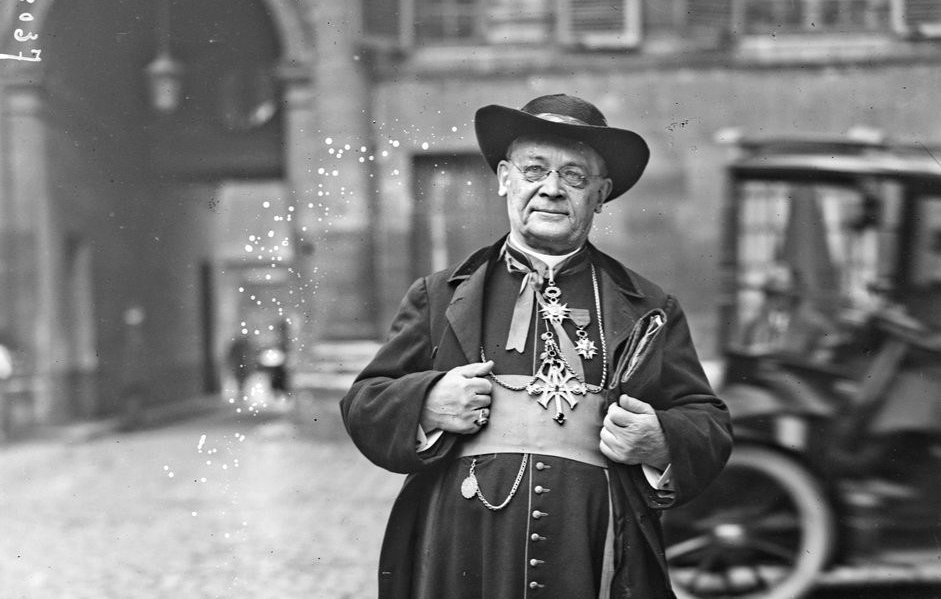 این تصویر یک مرد با لباس‌های سنتی و رسمی را نمایش می‌دهد. او کلاهی بزرگ بر سر دارد و لباسش بلند و مشکی است که با دکمه‌های درخشانی تزئین شده است. مرد در حال ایستادن بر روی سنگ‌فرش خیابانی است و در پس‌زمینه، ساختمان‌های قدیمی و یک خودروی تاریخی به چشم می‌خورد. به نظر می‌رسد او شخصیتی مهم یا مذهبی باشد. نور و کنتراست تصویر موجب برجسته شدن جزئیات لباس و چهره‌اش شده است.