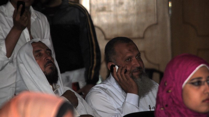 L'image montre des hommes et des femmes dans un environnement qui semble être un rassemblement ou une conférence. Deux hommes au premier plan portent des vêtements traditionnels, l'un d'eux est exprimé en train de passer un coup de téléphone. L'arrière-plan montre d'autres personnes, certaines concentrées sur l'événement, et des couleurs variées dans les vêtements, y compris une femme portant un hijab rose. L'atmosphère semble sérieuse et attentive.