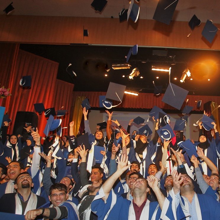 تظهر الصورة مجموعة كبيرة من الطلاب في حفل تخرج، وهم يرتدون قبعات التخرج والعباءات التقليدية. يبدو الجميع في حالة من الفرح والسعادة، حيث يرفعون قبعاتهم في الهواء احتفالاً بالإنجاز. الخلفية مليئة بالزينة، وهناك أضواء مسلطة تعزز من أجواء الحفل. تعكس الصورة روح الفخر والنجاح في هذه المناسبة المهمة في حياة الطلاب.