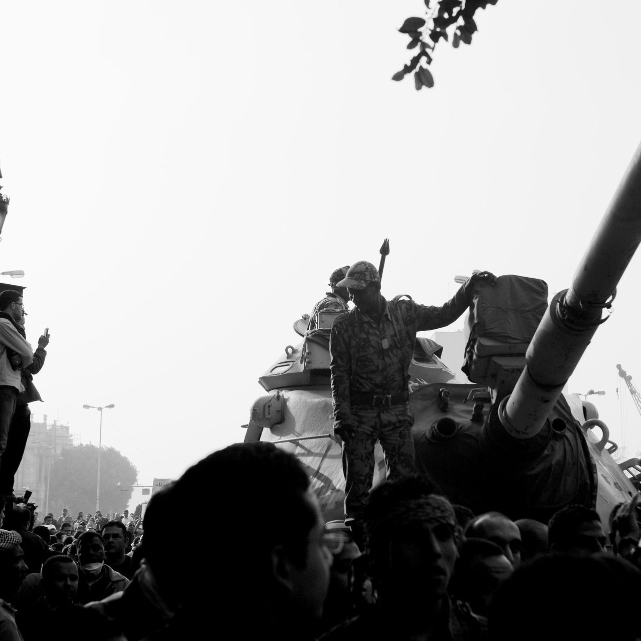 The image depicts a scene of intense activity, likely in an urban setting. In the foreground, a soldier stands on a tank, raising an arm, possibly in a gesture of authority or encouragement. The background is filled with a dense crowd, indicating a significant public event, possibly a protest or military presence. The overall tone of the image is stark, being presented in black and white, which adds to its dramatic effect. The atmosphere seems charged with emotion and energy, with some individuals observing or participating actively in the scene.