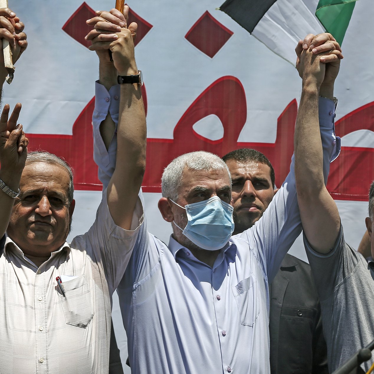 الصورة تُظهر مجموعة من الرجال في تجمع حاشد، حيث يرفع بعضهم أيديهم في إشارة للتضامن أو الاحتجاج. يرتدي أحد الأشخاص في المنتصف قناعاً للوجه، مما يشير إلى أنه قد يكون في سياق صحي معين. خلفهم يوجد لافتة كبيرة مكتوب عليها نص باللون الأحمر. الجو العام يوحي بالتعبير عن موقف أو رسالة معينة.