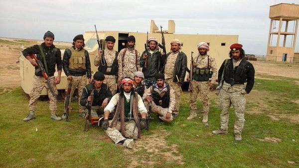 L'image montre un groupe de personnes en tenue militaire, armées et posant devant un véhicule. Ils semblent être sur un terrain en extérieur, peut-être dans un environnement désertique ou rural. Les hommes affichent diverses expressions et postures, certains sont debout tandis que d'autres sont assis. Leurs vêtements et accessoires suggèrent qu'ils font partie d'une unité militaire ou paramilitaire. En arrière-plan, on aperçoit un bâtiment et une tour d'eau.