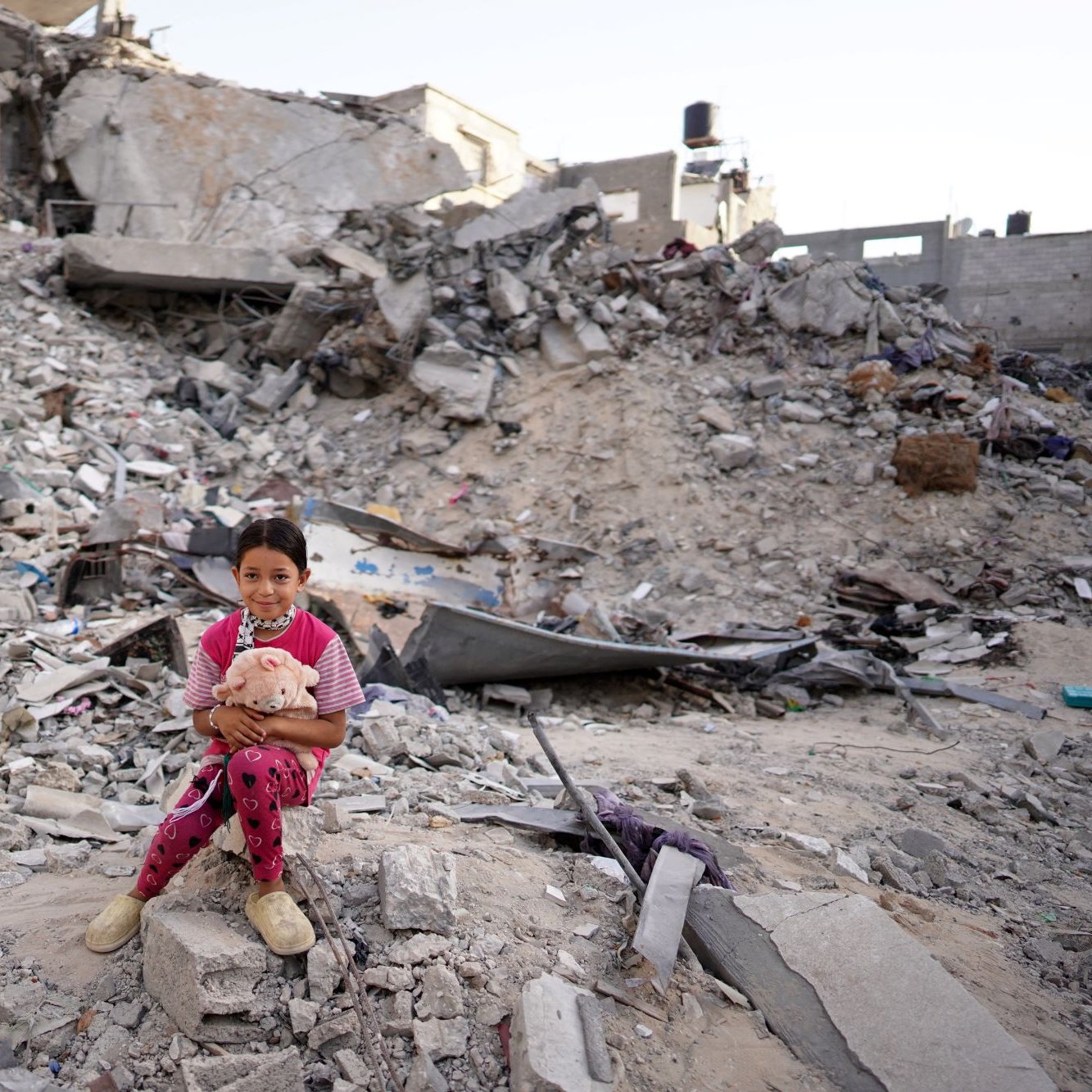 در این تصویر، دخترکی کوچک بر روی زمین در میان ویرانه‌های یک ساختمان نشسته است. در پس‌زمینه، تخریب‌های وسیع و انبوهی از آجرها و مصالح ساختمانی دیده می‌شود. دخترک با لباسی صورتی و شلوار آبی، عروسکی در آغوش دارد و به دوربین نگاه می‌کند. وضعیت اطراف او به وضوح نشان‌دهنده ویرانی و داغی ناشی از یک حادثه یا جنگ است. احساساتی از معصومیت و ناامیدی در این تصویر قابل مشاهده است.