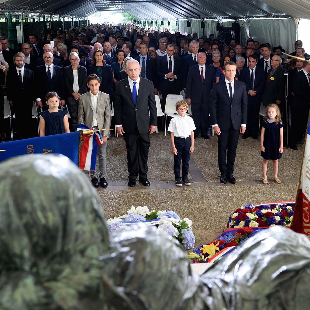 L'image montre une cérémonie commémorative avec un grand nombre de personnes rassemblées sous une tente. On peut voir des adultes en costumes et des enfants bien habillés. Au centre, deux hommes, dont l'un semble être un dirigeant, sont debout, les mains sur le cœur, en signe de respect. Il y a également des drapeaux et des gerbes de fleurs autour d'eux. L'ambiance semble solennelle et respectueuse, avec une attention particulière portée aux éléments de mémoire et d'honneur.