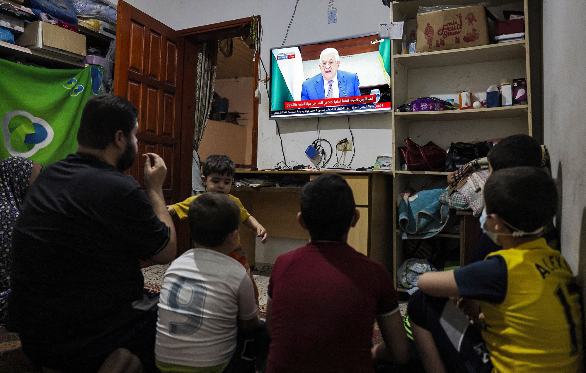 L'immagine mostra un gruppo di bambini seduti a terra, rivolti verso un televisore che trasmette un programma. Un adulto, probabilmente un genitore o un parente, è accanto a loro. La stanza sembra essere piccola e modesta, con mobili semplici e un'atmosfera informale. Sullo schermo si vede un uomo che parla, probabilmente un leader o un personaggio pubblico. I bambini appaiono curiosi e coinvolti nella visione.