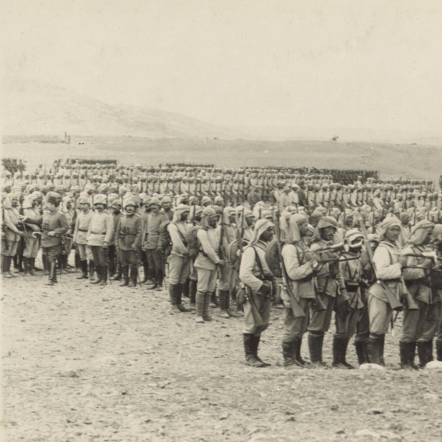 The image depicts a large group of soldiers lined up in a formation during a military event, likely a parade or review. The soldiers are dressed in uniforms typical of early 20th-century military attire, complete with helmets and boots. Some are holding rifles, and there are banners or flags present. In the background, more soldiers can be seen in organized rows, suggesting a significant military presence. The setting appears to be an open field, with hills or mountains visible in the distance, indicating a potentially historic or ceremonial occasion. The image has a monochromatic tone, typical of photographs from that era.