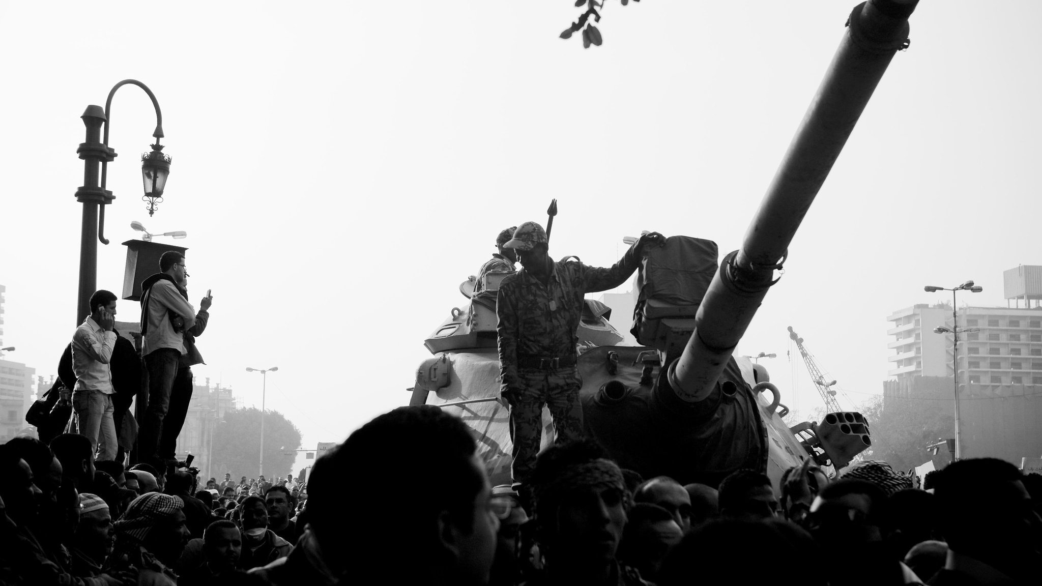 L'image montre une scène de manifestation avec un tank au premier plan. Des soldats sont visibles, dont un se tient debout sur le tank, levant un bras en signe de protestation ou de triomphe. Au premier plan, on aperçoit une foule de manifestants, certains prenant des photos ou filmant la scène. L'atmosphère est tendue, peut-être en raison de la situation politique. L'image est présentée en noir et blanc, ce qui renforce le côté dramatique de la scène.