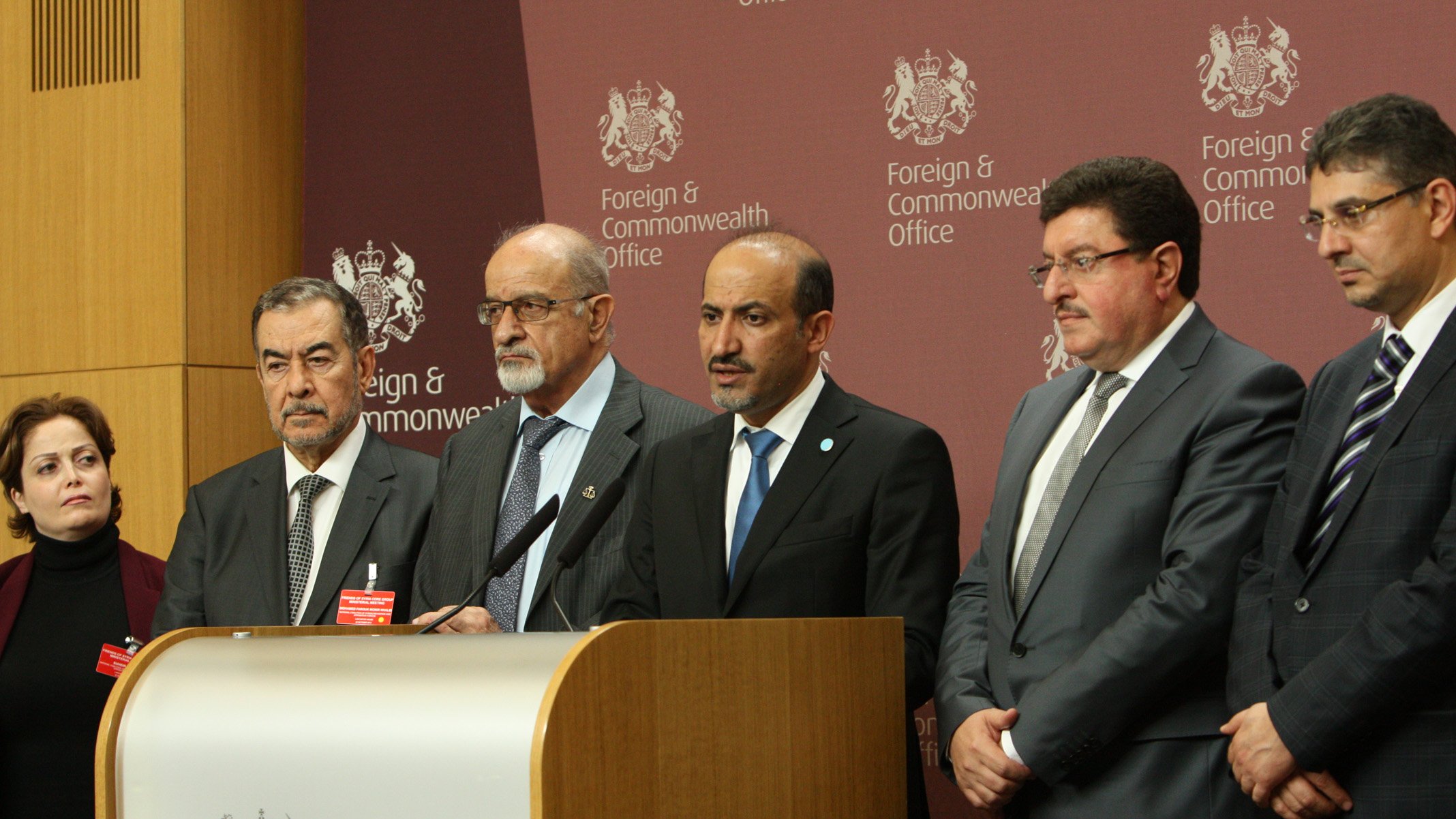 L'image montre un groupe de plusieurs personnes lors d'une conférence de presse. Elles se tiennent devant un podium avec un emblème officiel, et une toile de fond affichant "Foreign & Commonwealth Office". Les individus paraissent sérieux et concentrés, probablement en train de faire une déclaration ou de répondre à des questions. Les personnes sont habillées de manière formelle, illustrant l'importance de l'événement.