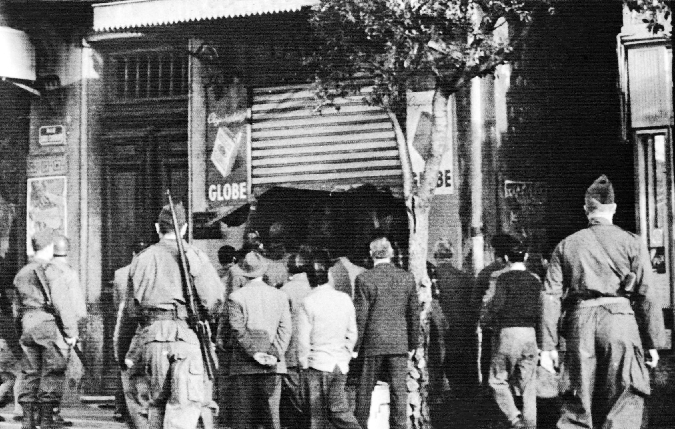 در این تصویر، گروهی از مردم در خیابان در حال نظارت به یک مکان هستند. تعدادی سرباز در نزدیکی آن‌ها ایستاده‌اند که به نظر می‌رسد در حال کنترل اوضاع هستند. این تصویر احتمالاً از یک زمان تاریخی گرفته شده است و نشان‌دهنده تنش یا رویدادی خاص در آن زمان است. حالت افراد و لباس‌هایشان به این احساس دامن می‌زند. برخی از افراد به سمت مغازه‌ای در حال حرکت هستند، در حالی که دیگران از دور نظاره‌گر هستند.