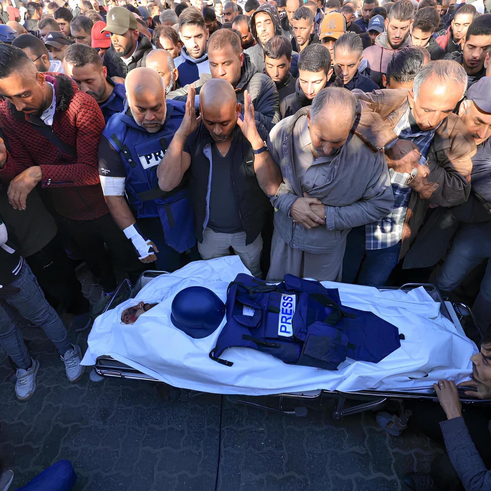 L'immagine mostra un gruppo di persone riunite in un'atmosfera di grande dolore e rispetto. Al centro, un corpo avvolto in un lenzuolo bianco è accompagnato da un giubbotto con la scritta "PRESS", suggerendo che si tratta di un giornalista. I presenti esprimono il loro cordoglio, alcuni con le mani giunte e altri con espressioni di tristezza. Il contesto sembra essere un evento commemorativo o funerario, evidenziando la gravità della situazione e il rispetto verso la persona deceduta.