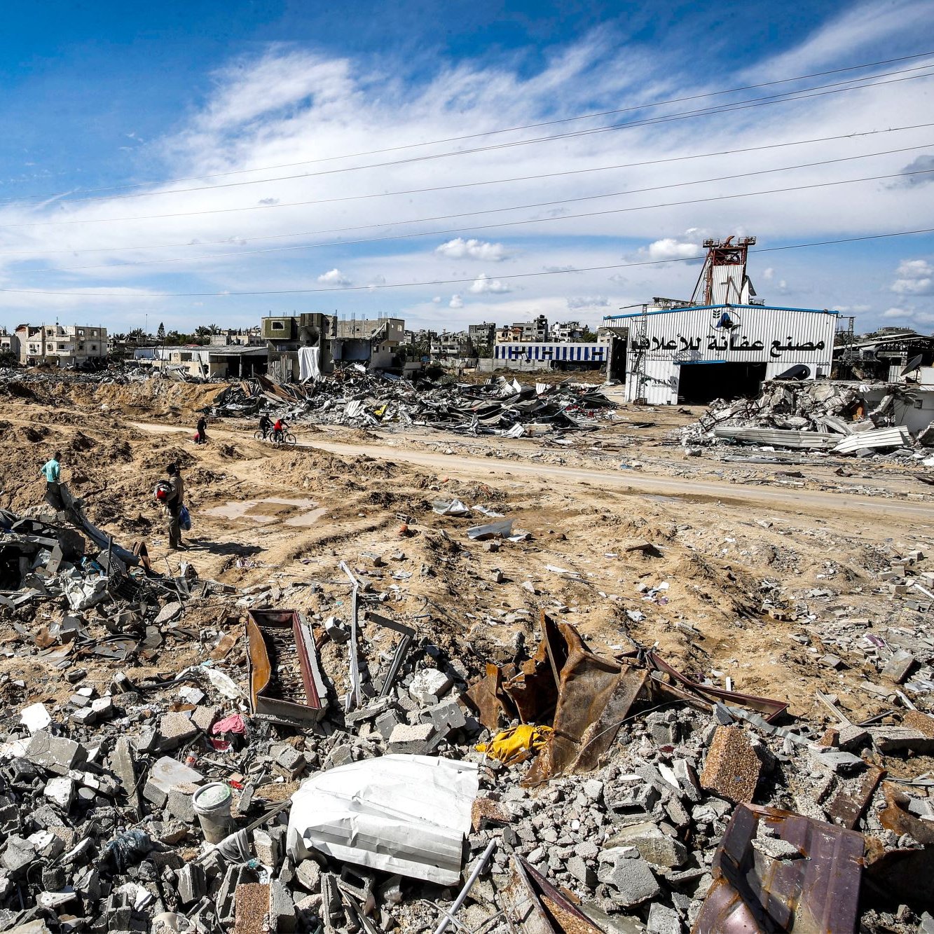 L'image montre un paysage dévasté, probablement dans une zone de conflit ou après une catastrophe. Le sol est jonché de débris, avec des morceaux de béton et de métal éparpillés. On peut apercevoir quelques personnes se déplaçant parmi les ruines, tandis qu'à l'arrière-plan, des bâtiments endommagés se dressent sous un ciel partiellement nuageux. Les lignes électriques sont visibles, indiquant une infrastructure perturbée. L'ambiance apparaît sombre et désolée, témoignant de la destruction qui a eu lieu.