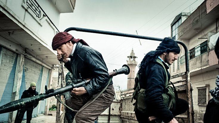 L'image montre des individus armés, apparemment dans un environnement urbain. Deux hommes sont en mouvement, l'un portant une arme lourde et l'autre un fusil. Ils semblent faire partie d'un groupe en action, probablement dans un contexte de conflit. L'arrière-plan montre des bâtiments et une ambiance sombre, qui renforcent le sentiment de tension. L'atmosphère est chargée et suggère un moment urgent.