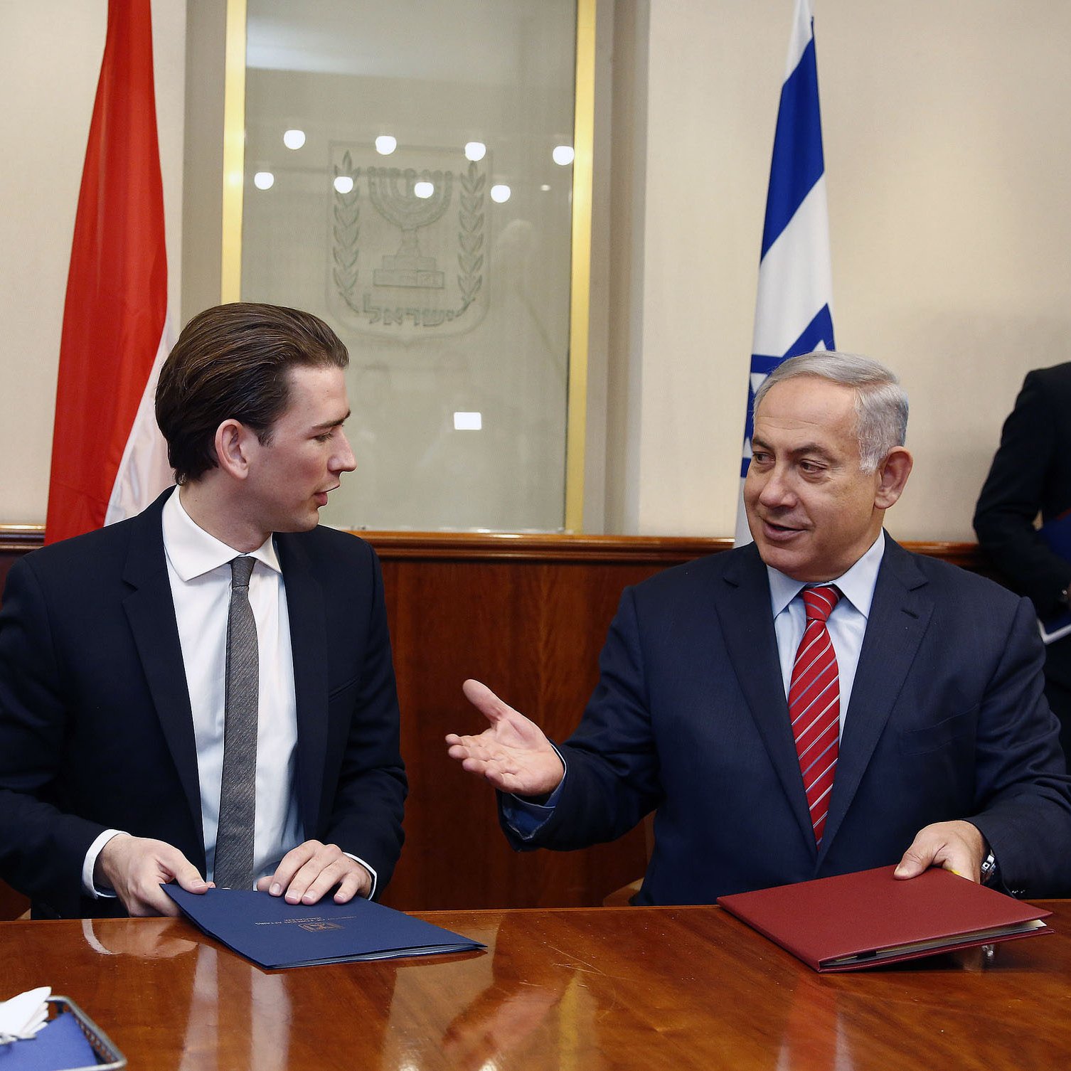 The image shows two men seated at a table in a formal setting. One man, wearing a black suit and tie, appears to be engaged in conversation with the other man, who is wearing a suit with a red tie. They are both holding folders and look to be discussing something important. In the background, there are flags visible, indicating a diplomatic context. The atmosphere appears professional and cooperative.