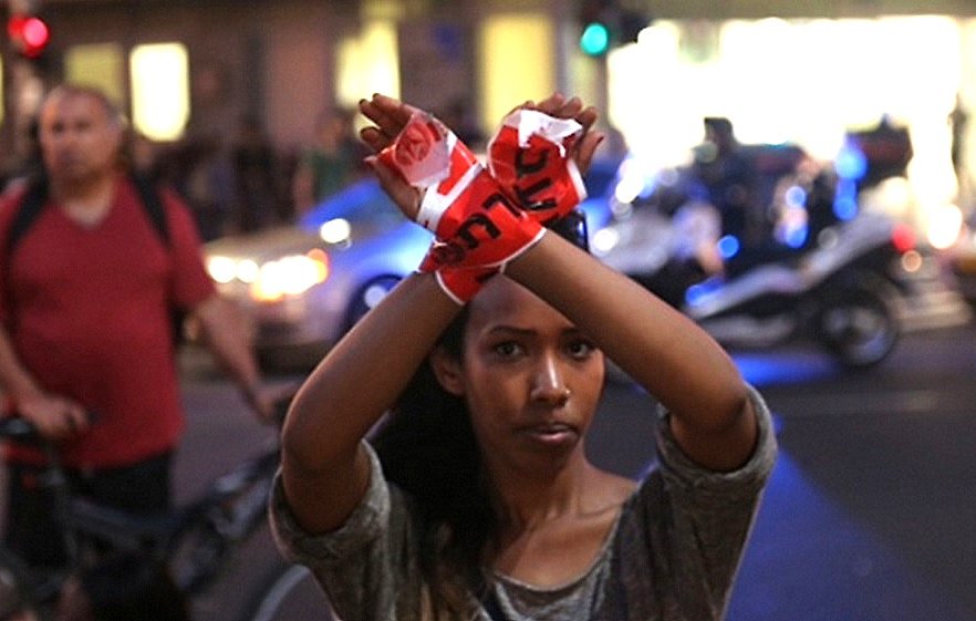 الصورة تظهر شابة ترفع يديها بطريقة تعبر عن الاحتجاج أو الاعتصام، حيث يتم ربط يديها بشريط يحمل كلمة أو شعار معين. الخلفية تحتوي على مشاهد حضرية، مع وجود أشخاص آخرين وسيارات تضيء بالأنوار. الأجواء تعكس لحظة من التوتر أو الحماسة، مما يدل على وجود حدث احتجاجي أو اجتماعي.