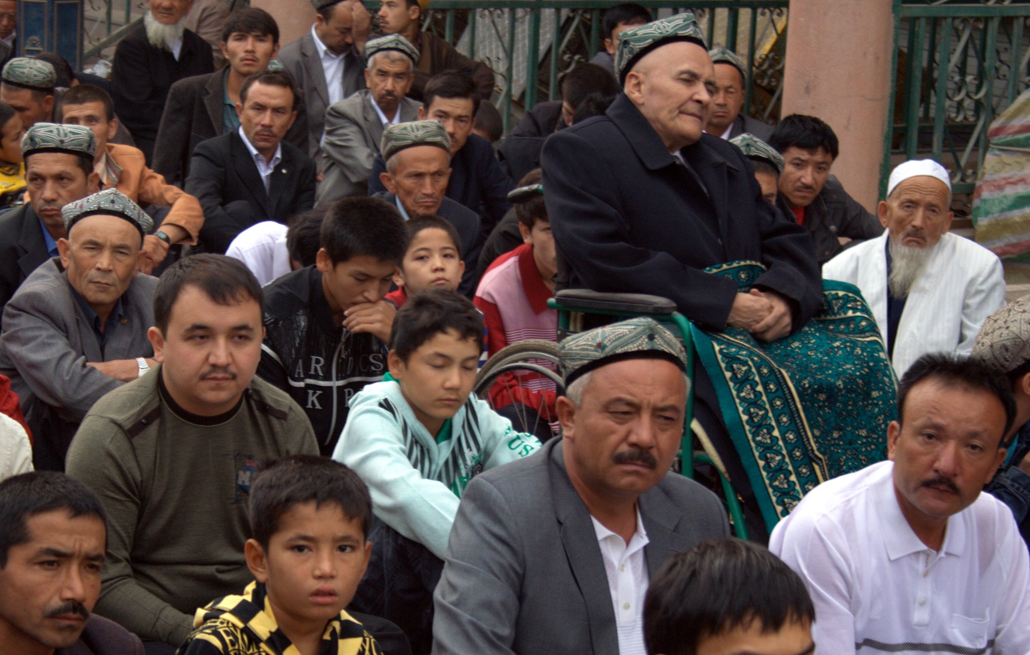 The image depicts a group of individuals seated together, primarily men, in a communal setting. They appear to be engaged in a gathering or possibly a religious ceremony, with some wearing traditional attire. In the foreground, there is an elderly man seated in a wheelchair, and others are attentively looking forward. The overall atmosphere reflects a sense of community and shared purpose. The individuals show a range of ages, from children to older men, indicating that the event likely holds significance for various generations.