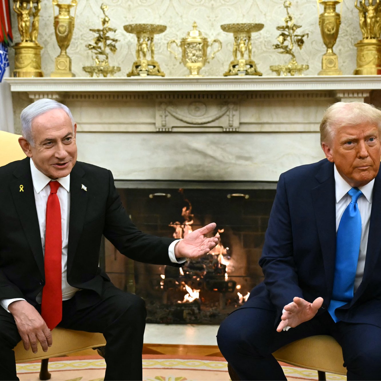 Nell'immagine si possono vedere due uomini seduti in poltrone di fronte a un camino. A sinistra c'è un uomo con una giacca nera, una cravatta rossa e un distintivo sul petto. A destra, l'altro uomo indossa un giubbotto blu e una cravatta rossa, anch'esso con un distintivo. Entrambi stanno parlando e sembrano impegnati in una conversazione. Sullo sfondo c'è un arredamento elegante, con decorazioni sulle pareti e un camino acceso.