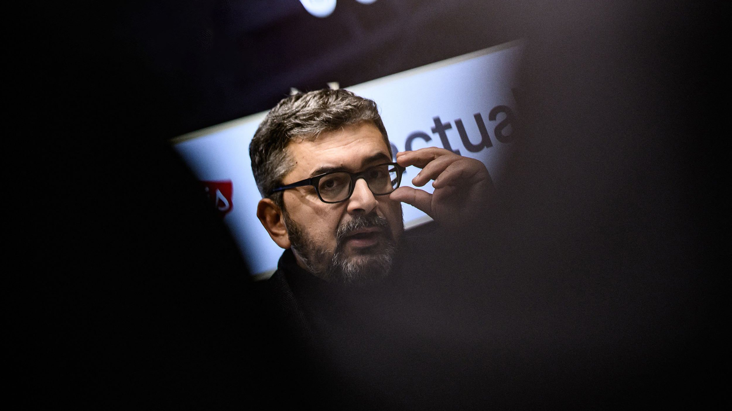 L'image montre un homme portant des lunettes et ayant une barbe courte. Il semble engagé dans une conversation ou une présentation, en touchant son visage avec une main. En arrière-plan, des éléments graphiques ou des logos sont flous, ce qui suggère qu'il se trouve dans un cadre professionnel ou public. L'éclairage met en avant son visage, tandis que des silhouettes floues d'autres personnes sont visibles autour.