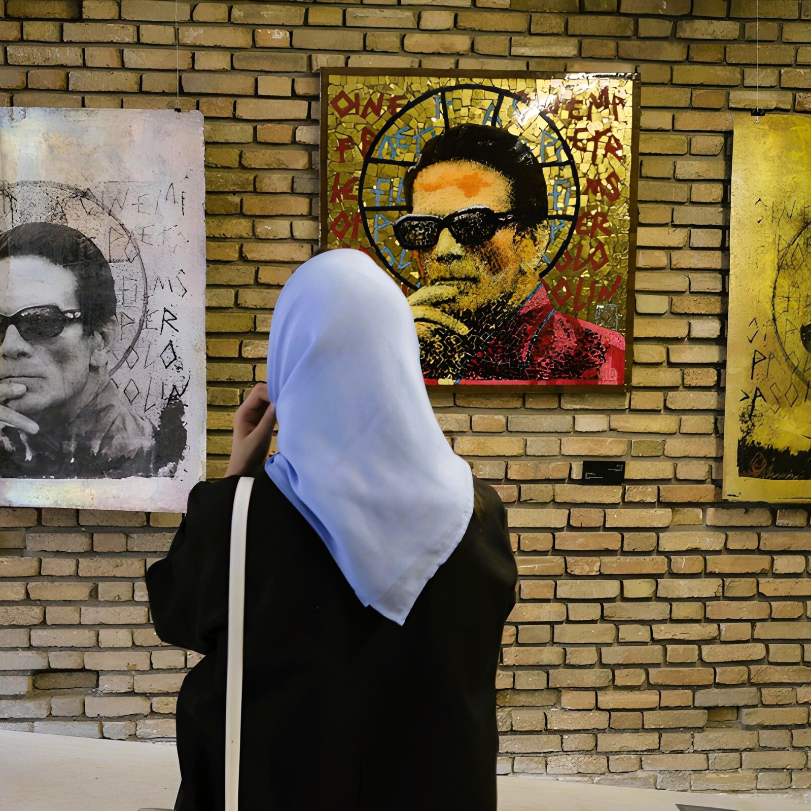 در تصویر، یک زن با پوشش اسلامی در حال تماشا به آثار هنری روی دیوار یک گالری است. این آثار شامل تصاویری از یک مرد با عینک آفتابی است که به نظر می‌رسد به صورت هنری و با رنگ‌های مختلف خلق شده‌اند. پس‌زمینه دیوار گالری از آجرهای قهوه‌ای رنگ ساخته شده است و فضای هنری خوبی را ایجاد کرده است. زن در حال تفکر و تماشا به این آثار هنری به نظر می‌رسد.