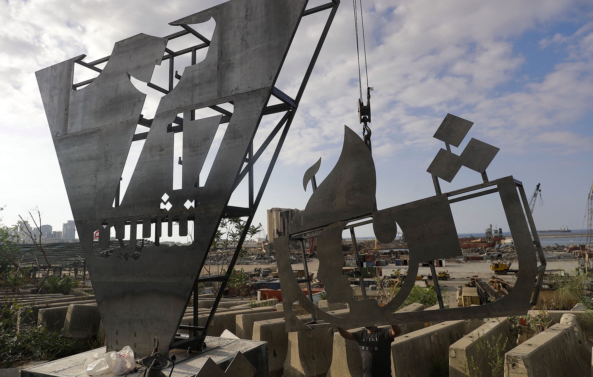 The image depicts two large, metallic sculptures or structures under construction or installation in an industrial outdoor setting. The sculptures show abstract forms, one resembling a stylized letter or shape and the other possibly depicting a ship or vessel. The background features a harbor with cranes and construction materials scattered around, suggesting an active worksite. The sky is partly cloudy, adding to the overall atmosphere of a busy environment.