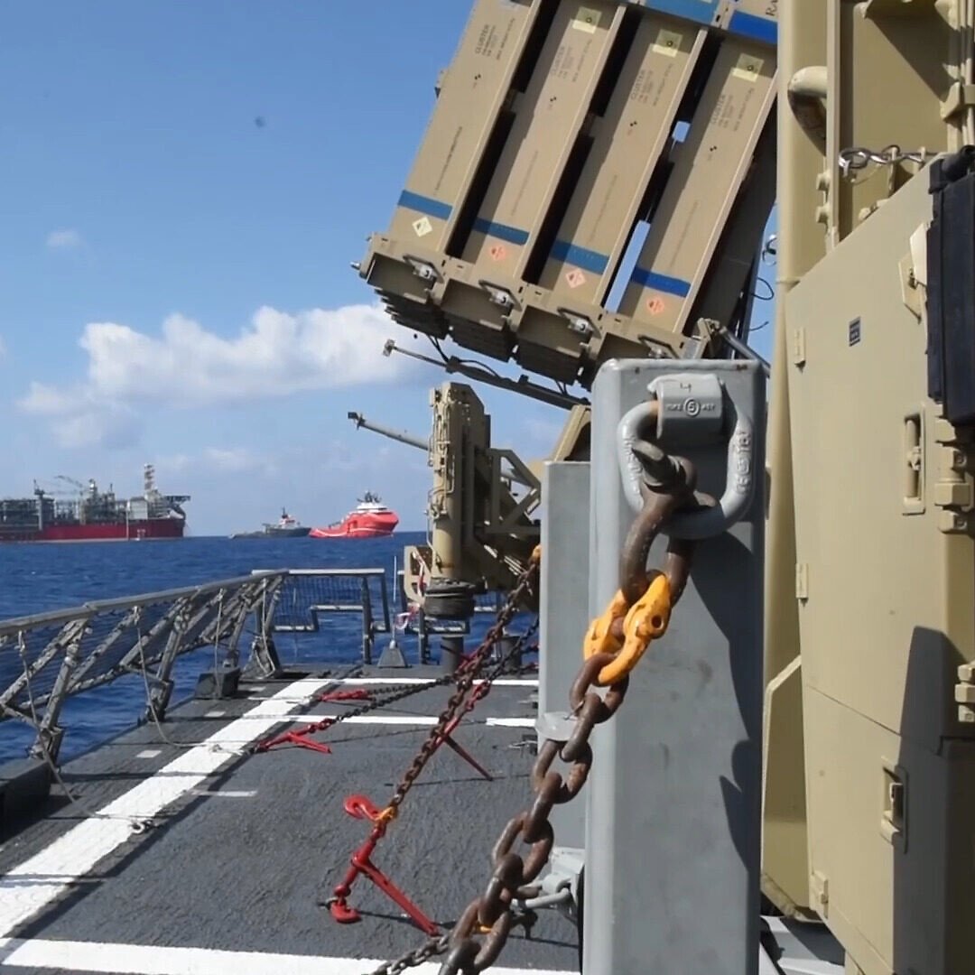 در تصویر یک نمای دریایی مشاهده می‌شود که شامل یک کشتی نظامی بخصوص با تسلیحات نصب شده بر روی آن است. در پس‌زمینه، چندین سکوی نفتی و کشتی دیگر در دریا دیده می‌شوند. آسمان آبی و روشن است و چند ابر نیز در آن وجود دارد. فضای کلی تصویر به نظر می‌رسد که در حال اجرای عملیات دریایی یا امنیتی است.