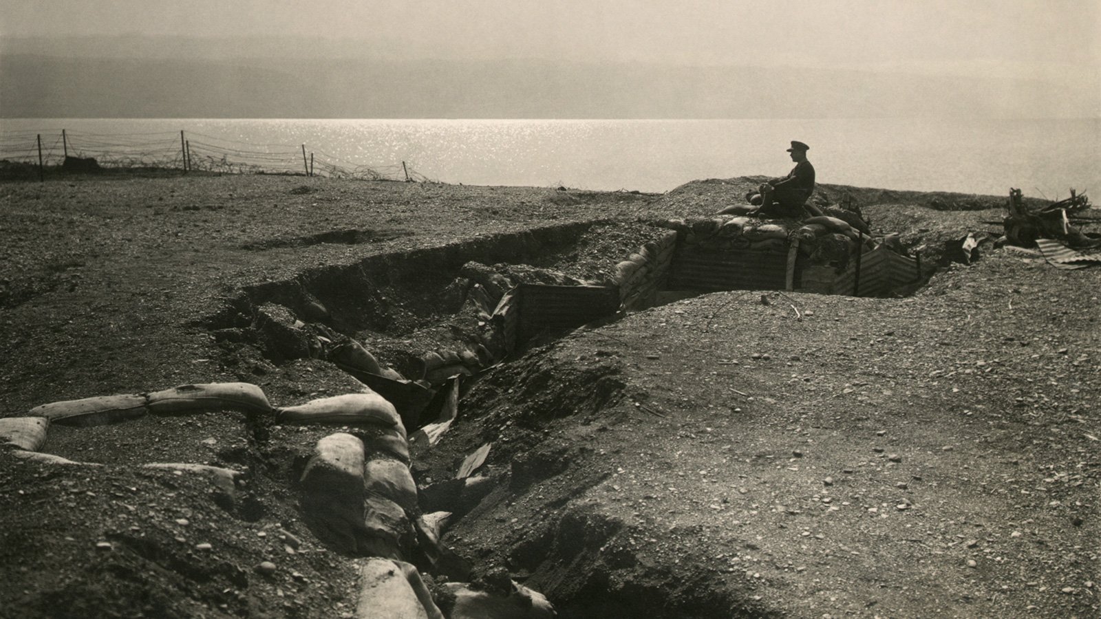 L'image représente un champ de bataille avec une tranchée. On peut voir un soldat assis sur les bords de la tranchée, qui semble surveiller les environs. Le terrain est accidenté et semé de sacs de sable, typiques des fortifications de la Première Guerre mondiale. À l'arrière-plan, on aperçoit une étendue d'eau qui pourrait être un lac ou une rivière. L'atmosphère générale est sombre et mélancolique, évoquant la dureté de la guerre.