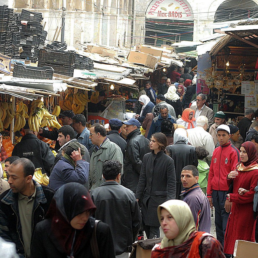 L'image montre un marché animé, probablement dans une ville du Moyen-Orient ou d'Afrique du Nord. Des stands sont remplis de fruits, notamment des bananes et des oranges. On peut voir de nombreuses personnes, vêtues de vêtements variés, se déplacer entre les étals. L'atmosphère est animée, avec des groupes discutant et des clients choisissant des marchandises. Des éclairages pendent du plafond, ajoutant une ambiance particulière au lieu.