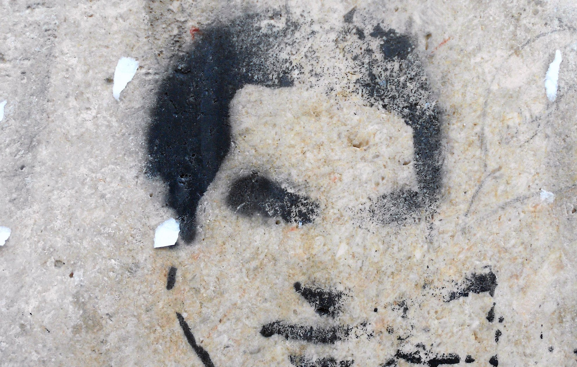 این تصویر شامل یک طرح یا گرافیتی روی دیوار است که چهره‌ای را به صورت سیاه و سفید نشان می‌دهد. جزئیات چهره به وضوح قابل تشخیص نیستند و بیشتر به صورت سایه‌ای به نظر می‌رسند. زمینه تصویر ممکن است از جنس بتن یا دیوار سنگی باشد که به مرور زمان دچار wear و تخریب شده است. 