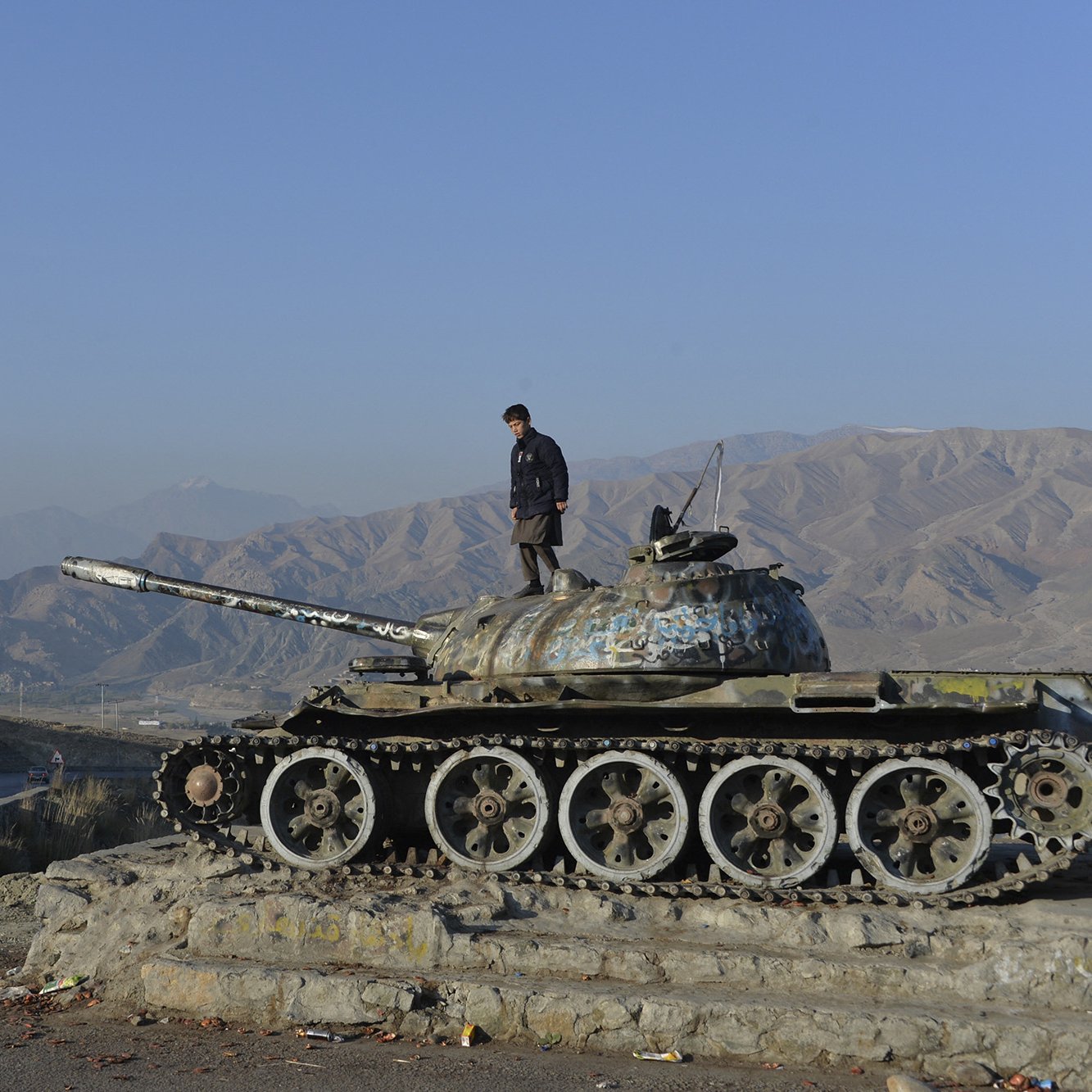 در این تصویر، یک تانک قدیمی با رنگ و زنگ‌زدگی مشاهده می‌شود که در یک نقطه مرتفع قرار دارد و فردی بر روی آن ایستاده است. پس‌زمینه شامل کوه‌های بلند و آسمان صاف است. در سمت راست تصویر، یک اتومبیل در حال حرکت در جاده‌ای کوهستانی دیده می‌شود. این صحنه نمایانگر تلاقی تاریخ و مناظر طبیعی است.