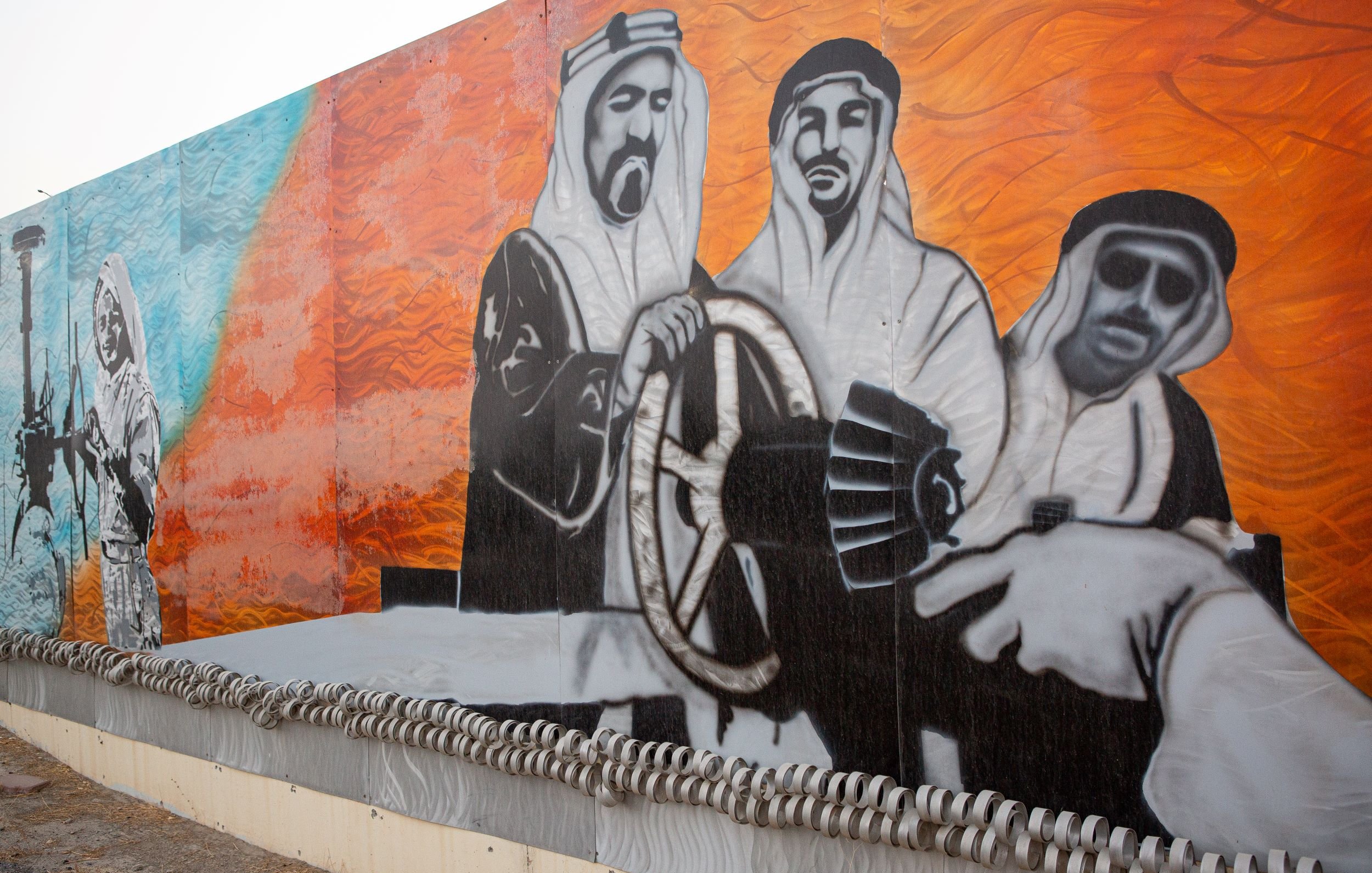 L'image représente une grande fresque murale. On y voit des figures en vêtements traditionnels arabes, probablement des hommes, qui semblent interagir avec des éléments symboliques liés à l'industrie pétrolière. L'arrière-plan est composé de couleurs vives, notamment des nuances de bleu et d'orange. Cette œuvre évoque des thèmes de culture, de tradition et de développement industriel, sur fond de paysages désertiques. La composition suggère un sens de la coopération et de l'engagement vers une vision commune.