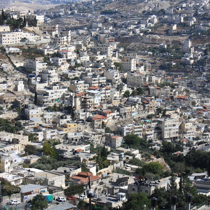L'image montre un paysage urbain densément construit avec de nombreuses maisons et bâtiments. On peut observer une variété d'architecture, allant des structures modernes aux constructions plus anciennes. Les collines environnantes sont également visibles, avec des zones verdoyantes et des routes serpentant à travers le paysage. La ville semble être située dans une région montagneuse, avec un éclairage naturel qui met en valeur les détails des bâtiments.