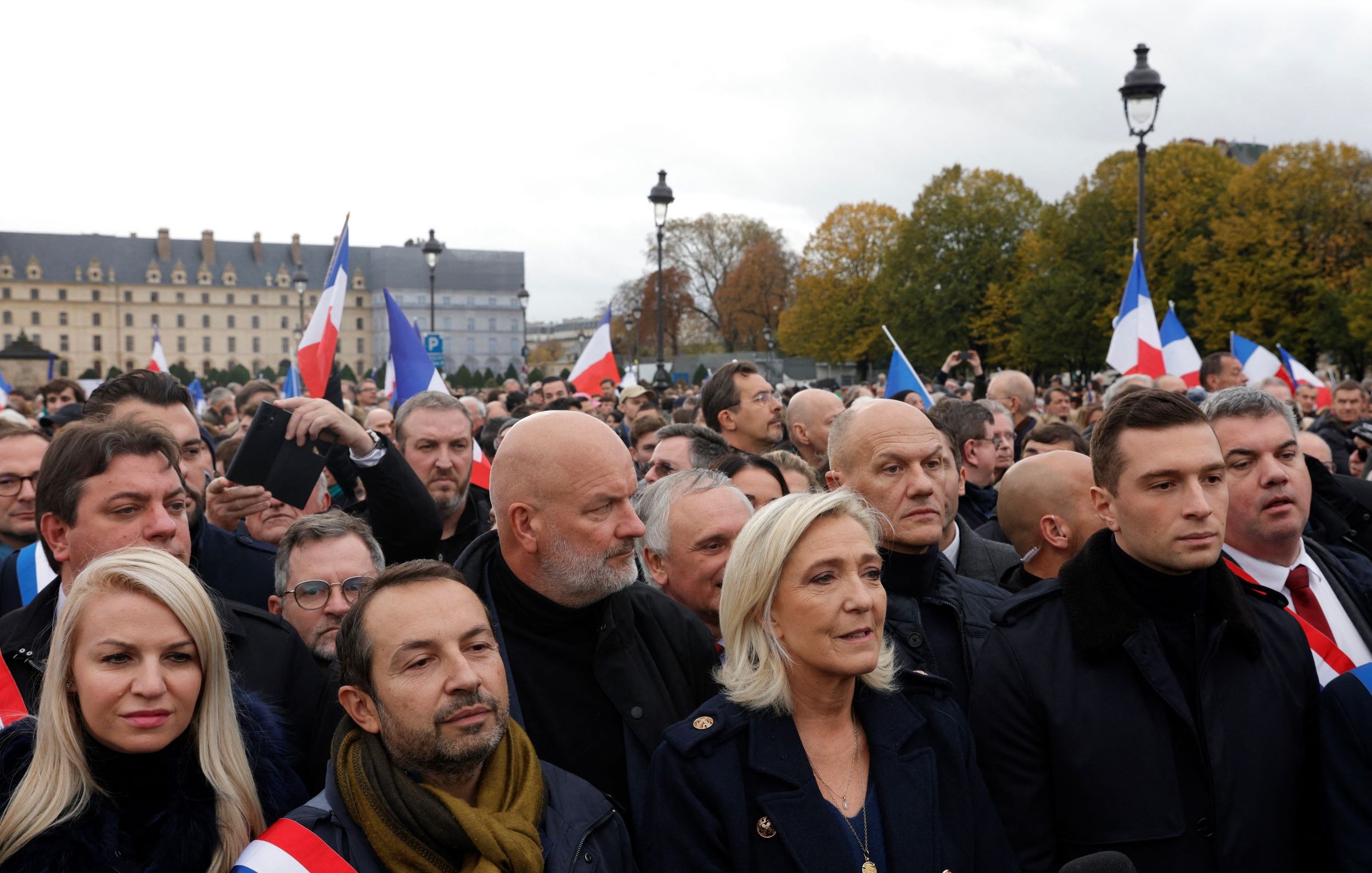 L'image montre une foule rassemblée lors d'un événement en plein air. Au premier plan, plusieurs personnes sont visibles, dont certaines portent des écharpes tricolores aux couleurs du drapeau français. On aperçoit également des drapeaux français agités par des membres de la foule. L'arrière-plan présente des bâtiments historiques et un ciel nuageux, ce qui donne une atmosphère de mobilisation. Les expressions des personnes semblent sérieuses et engagées, témoignant de l'importance de l'événement.