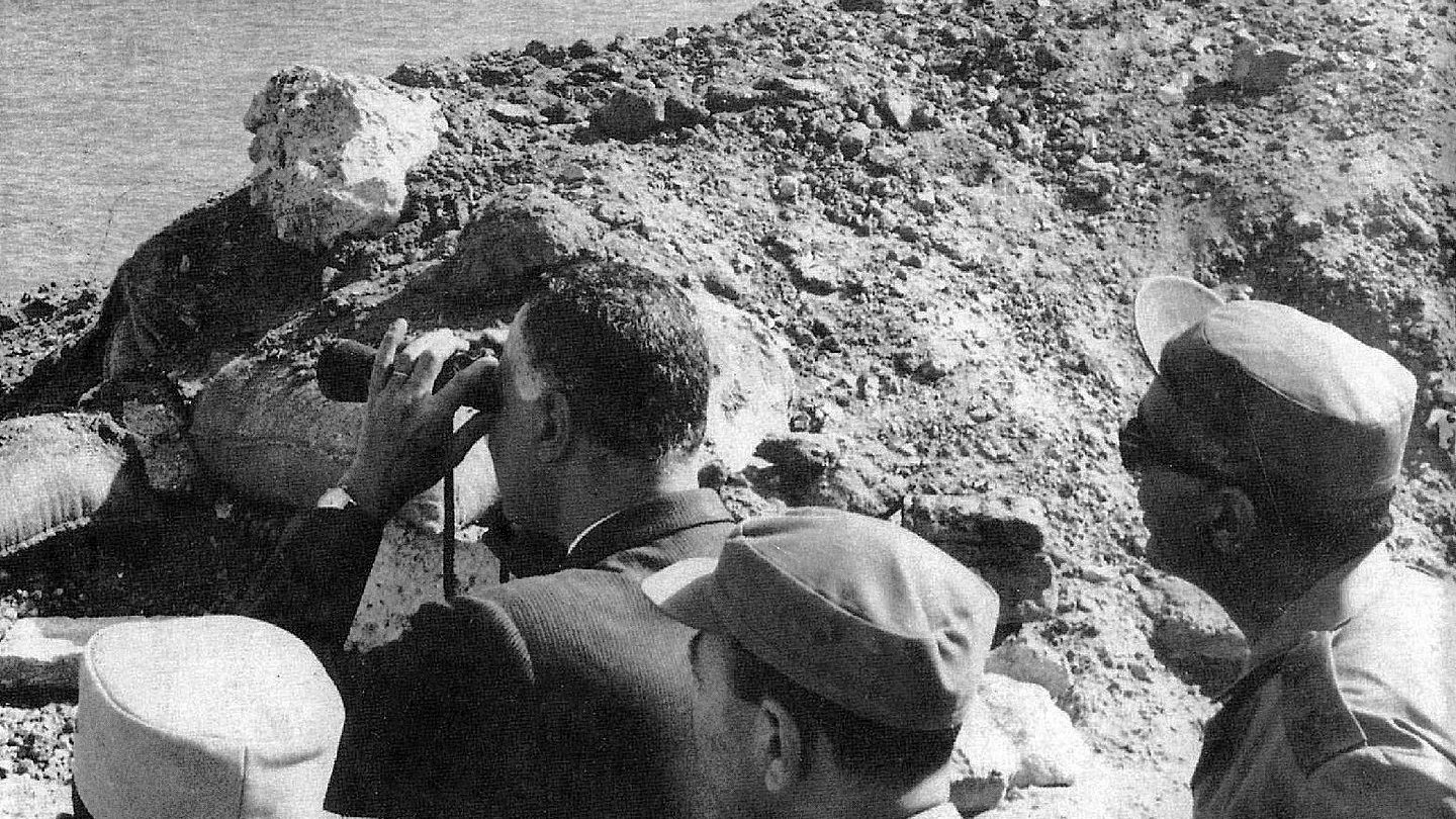 L'image montre un groupe d'hommes, dont l'un semble observer quelque chose au loin à l'aide de jumelles. Ils se tiennent près d'une rive d'eau, probablement à proximité d'un site de construction ou d'une digue, entourés de débris ou de terre. L'environnement semble suggérer une période historique, peut-être liée à des événements militaires ou politiques. Les hommes portent des uniformes ou des vêtements formels, ce qui indique qu'ils occupent probablement des positions officielles.