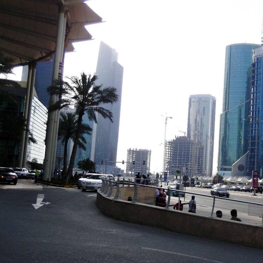 تظهر الصورة مشهداً حضرياً لمدينة حديثة، حيث تتداخل المباني الشاهقة مع تشكيلة متنوعة من التصاميم المعمارية. في الجهة اليسرى، توجد أشجار النخيل التي تضيف لمسة طبيعية على المشهد. يتواجد على الطريق مجموعة من السيارات، مما يشير إلى حركة المرور في المنطقة. السماء تظهر بلون فاتح، مما يوحي بأنها يوم مشمس. العمارات تتنوع في ارتفاعاتها وأشكالها، مما يعكس الطابع العصري للمدينة.