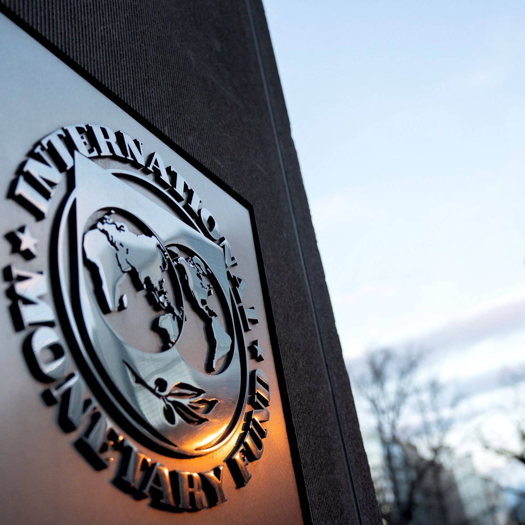 La imagen muestra el emblema del Fondo Monetario Internacional (FMI) en primer plano. El logo presenta un diseño que representa un globo terráqueo rodeado de hojas, simbolizando la cooperación internacional y el desarrollo económico. De fondo, se puede ver un edificio moderno con una fachada de cristal y un cielo parcialmente nublado, lo que sugiere un ambiente urbano y contemporáneo.