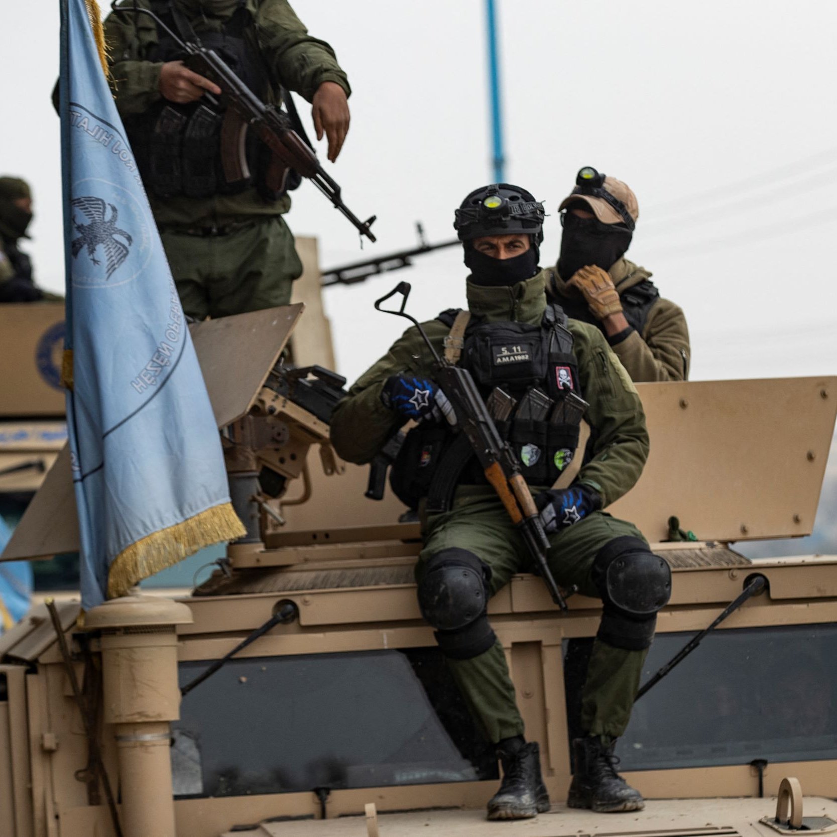 تظهر في الصورة مجموعة من الجنود العسكريين أثناء وجودهم في مركبة مدرعة. يرتدي الجنود زيًا عسكريًا ويظهر على بعضهم معدات تكتيكية وأسلحة. أحد الجنود يجلس على الجزء العلوي من المركبة، بينما يظهر آخرون يقفون عليها. هناك علم أزرق يعود لجهة معينة يبدو أنه متعلق بمهام حفظ السلام. الأجواء المحيطة تبدو حضرية مع وجود أشجار ومرافق عامة في الخلفية.