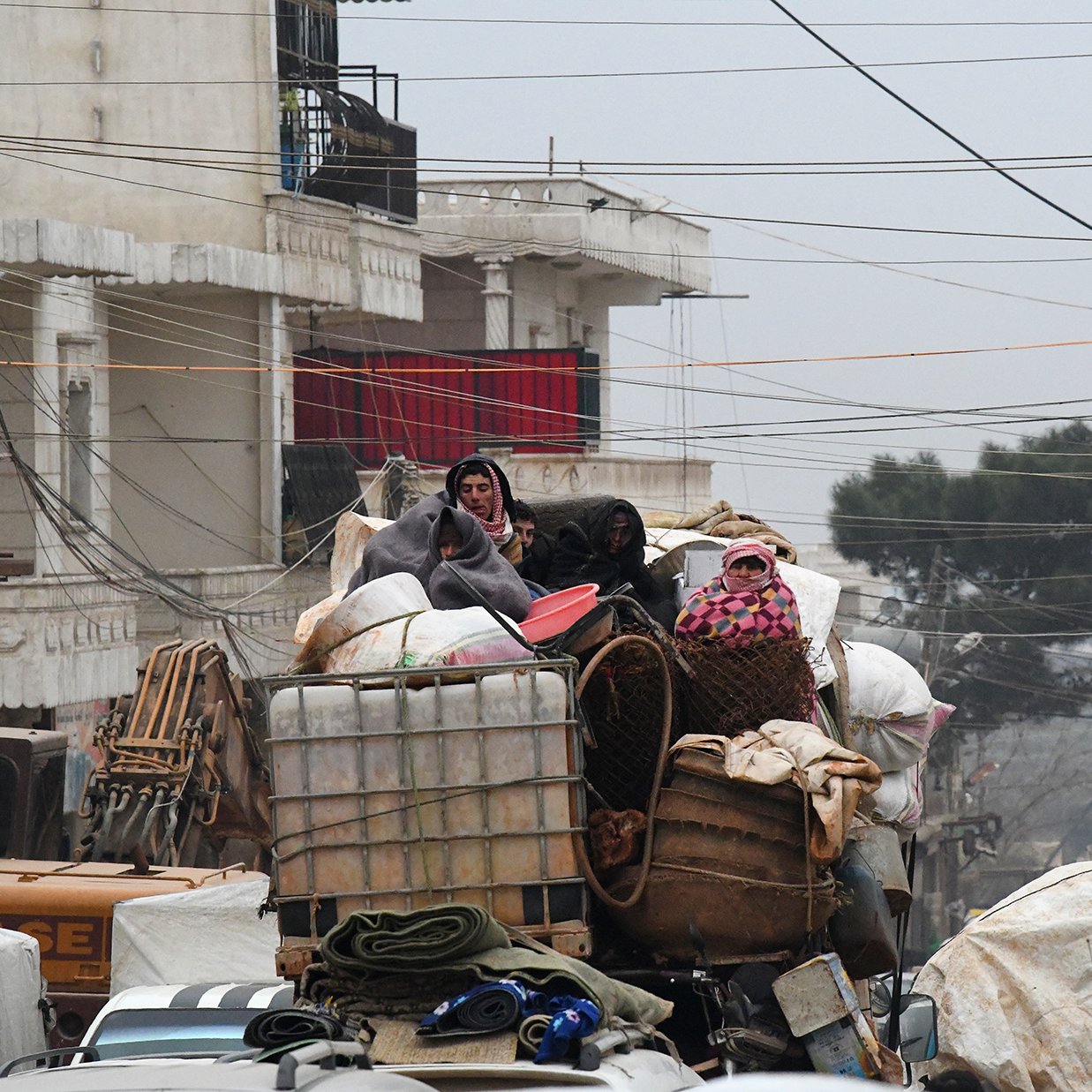 در این تصویر، گروهی از افراد در حال سفر بر روی یک کامیون بار هستند. آن‌ها به طور واضحی در میان بار زیادی از محصولات و وسایل قرار دارند. برخی از افراد پوشیده از لباس‌های ضخیم و شال‌های رنگی به نظر می‌رسند که به دلیل سرما به خود پیچیده‌اند. محیط اطراف شلوغ و شهرنشین به نظر می‌رسد، با ساختمان‌ها و خطوط برق مشخص. وضعیت کلی تصویر احساس چالش و سختی را القا می‌کند.
