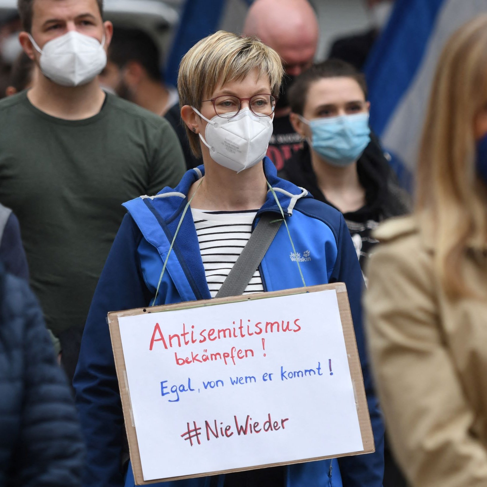 تظهر الصورة مجموعة من الأشخاص في مظاهرة، حيث يرتدي الجميع أقنعة واقية. في المقدمة، توجد امرأة تحمل لافتة مكتوب عليها "كفى من معاداة السامية!" باللغة الألمانية، بينما تشير العبارة الأخرى على اللافتة إلى "لا للإعادة!"، مما يعكس موقفًا معارضًا للتمييز. البيئة المحيطة تبدو مشغولة بعدد من المتظاهرين الآخرين، مما يدل على وجود حشد من الأشخاص خلال هذا الحدث.