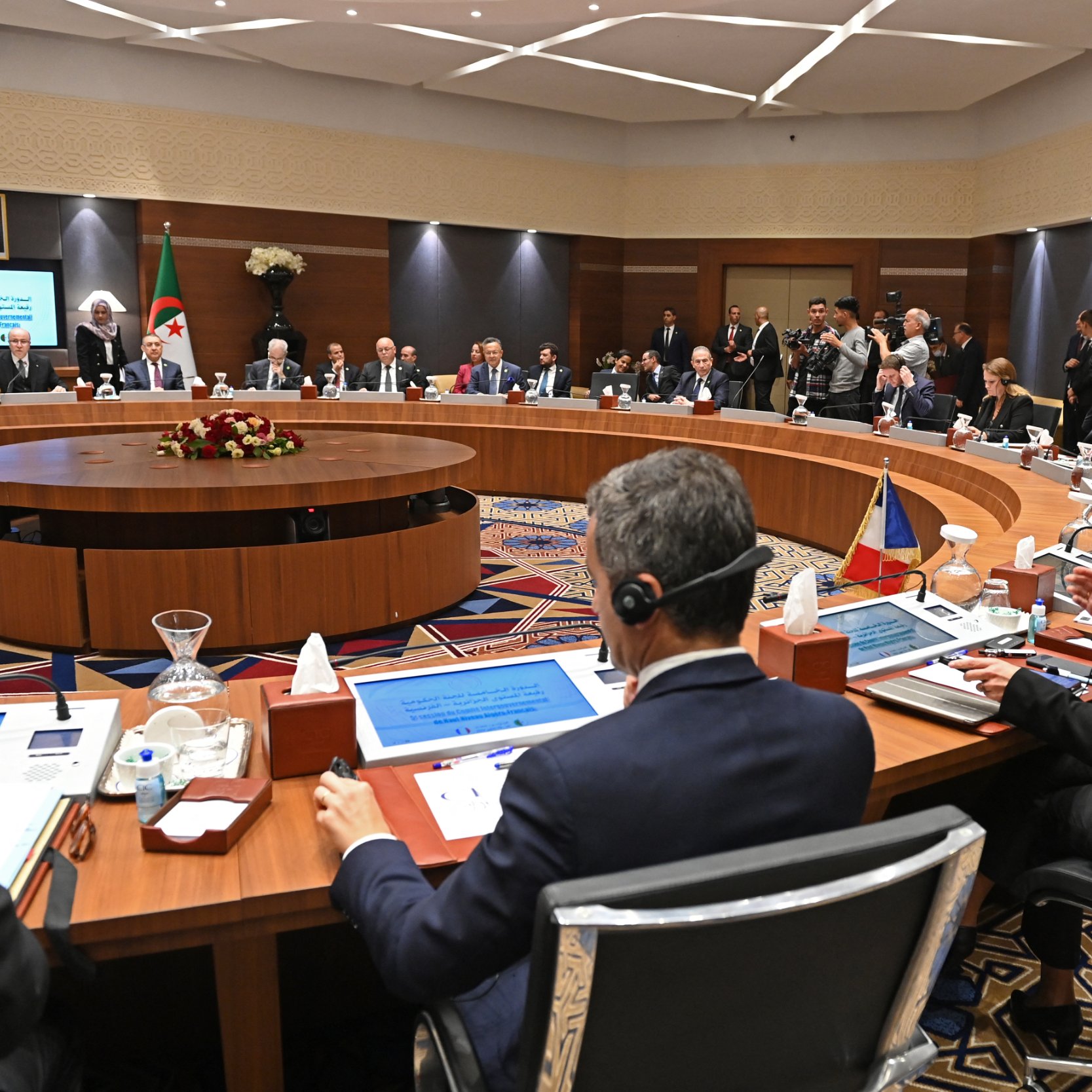 L'image montre une réunion formelle dans une salle de conférence. De nombreuses personnes sont assises autour d'une grande table ovale, probablement des représentants de différents pays ou organisations. Certains participants portent des casques d'écoute, indiquant qu'il pourrait y avoir des traductions simultanées. La salle est décorée avec des fleurs et des éléments modernes, et il y a des écrans affichant des informations pertinentes. Certaines personnes se tiennent debout en arrière-plan, observant les discussions. L'atmosphère semble professionnelle et concentrée.