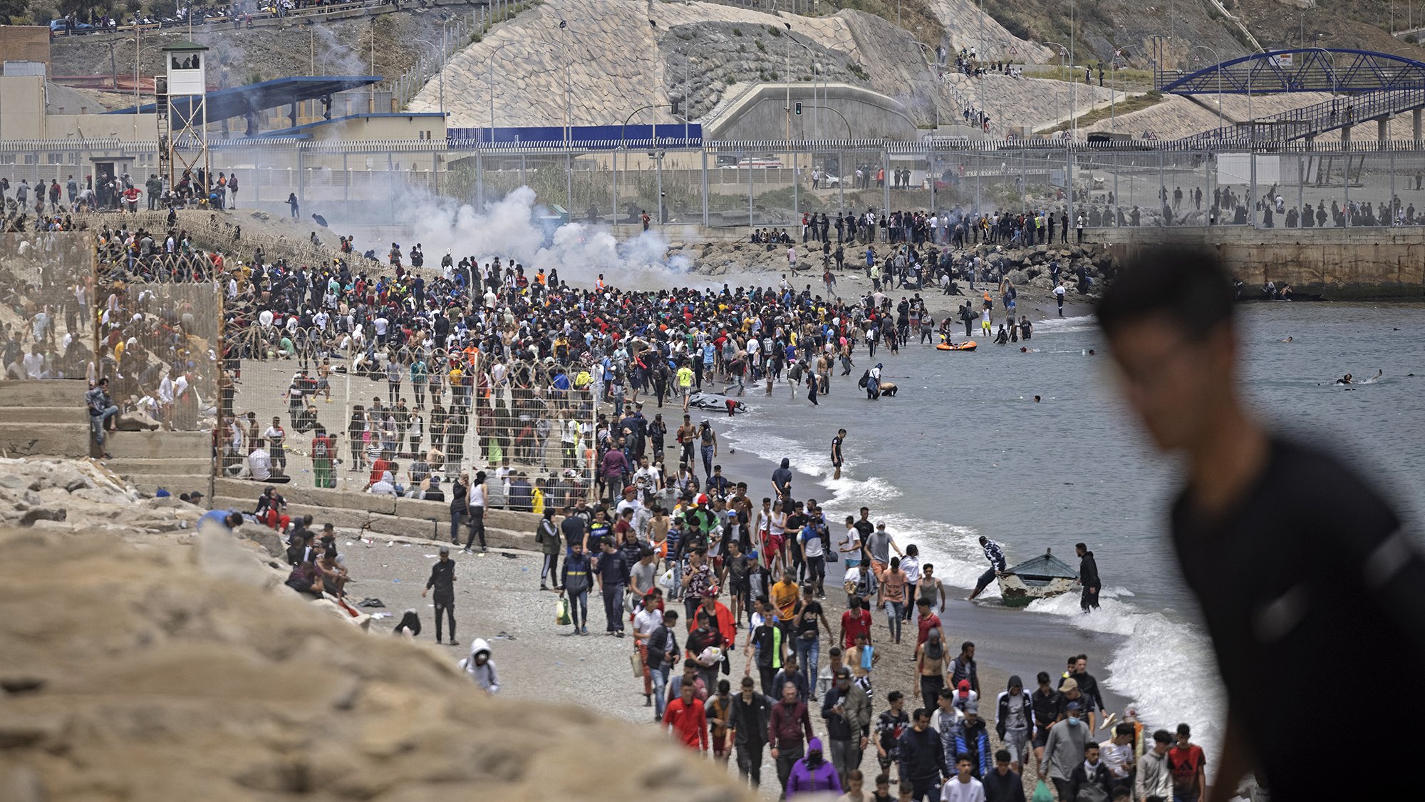 L'image montre une foule nombreuse sur une plage, avec des personnes rassemblées près de l'eau et d'autres sur le rivage. En arrière-plan, on peut voir des collines ainsi que des structures de béton. Il y a également des éléments de chaos, comme de la fumée, ce qui pourrait indiquer une situation tendue ou un événement significatif. Des gens semblent se déplacer, certains en direction de la mer, tandis que d'autres se trouvent sur le sable ou dans l'eau.