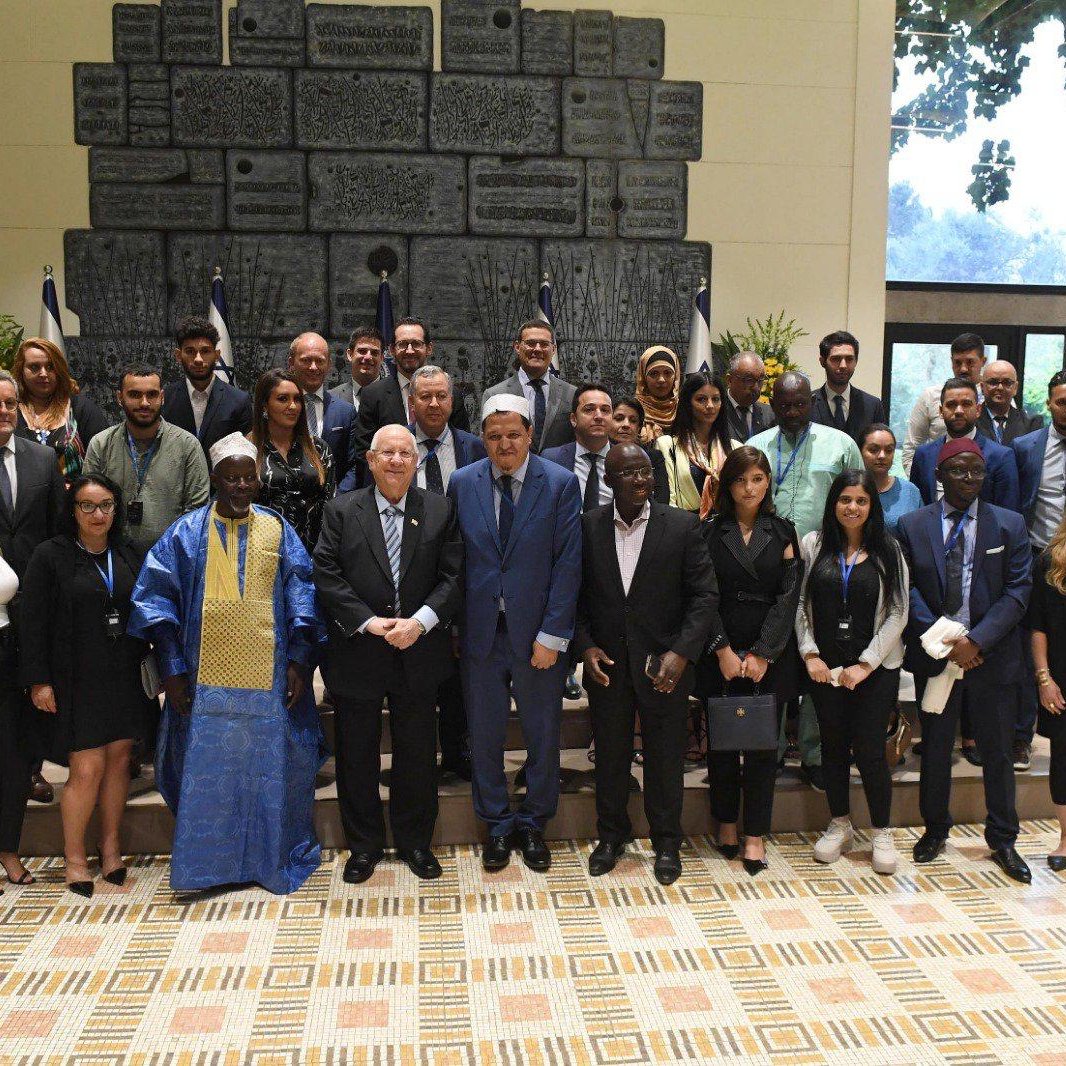 La imagen muestra a un grupo de personas posando juntas en un espacio interior bien iluminado. Al fondo, se pueden ver banderas y una pared decorativa. El grupo incluye hombres y mujeres de diversas apariencias, algunos con trajes formales y otros con atuendos tradicionales. Todos están sonriendo y parecen reunidos para una ocasión especial, posiblemente un evento diplomático o cultural. La disposición del grupo sugiere una atmósfera de cooperación y celebración.