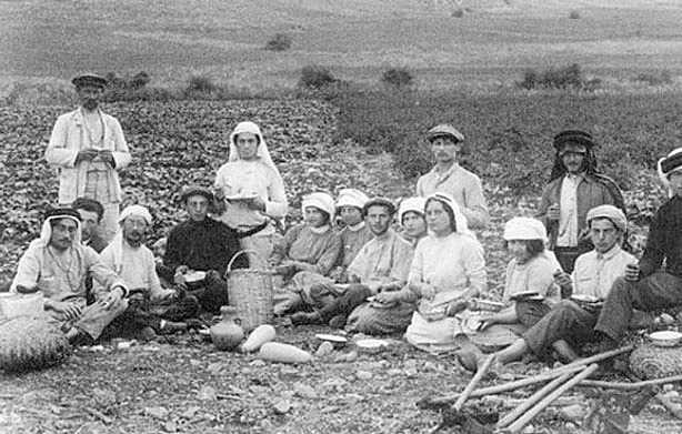 The image depicts a group of people gathered in an agricultural setting, likely in the early 20th century based on the clothing styles. There are both men and women, some wearing traditional garments and hats. They appear to be taking a break from farming activities, sitting on the ground with various tools and baskets nearby. The landscape in the background features fields and hills, suggesting a rural environment. Overall, it captures a moment of community among laborers.